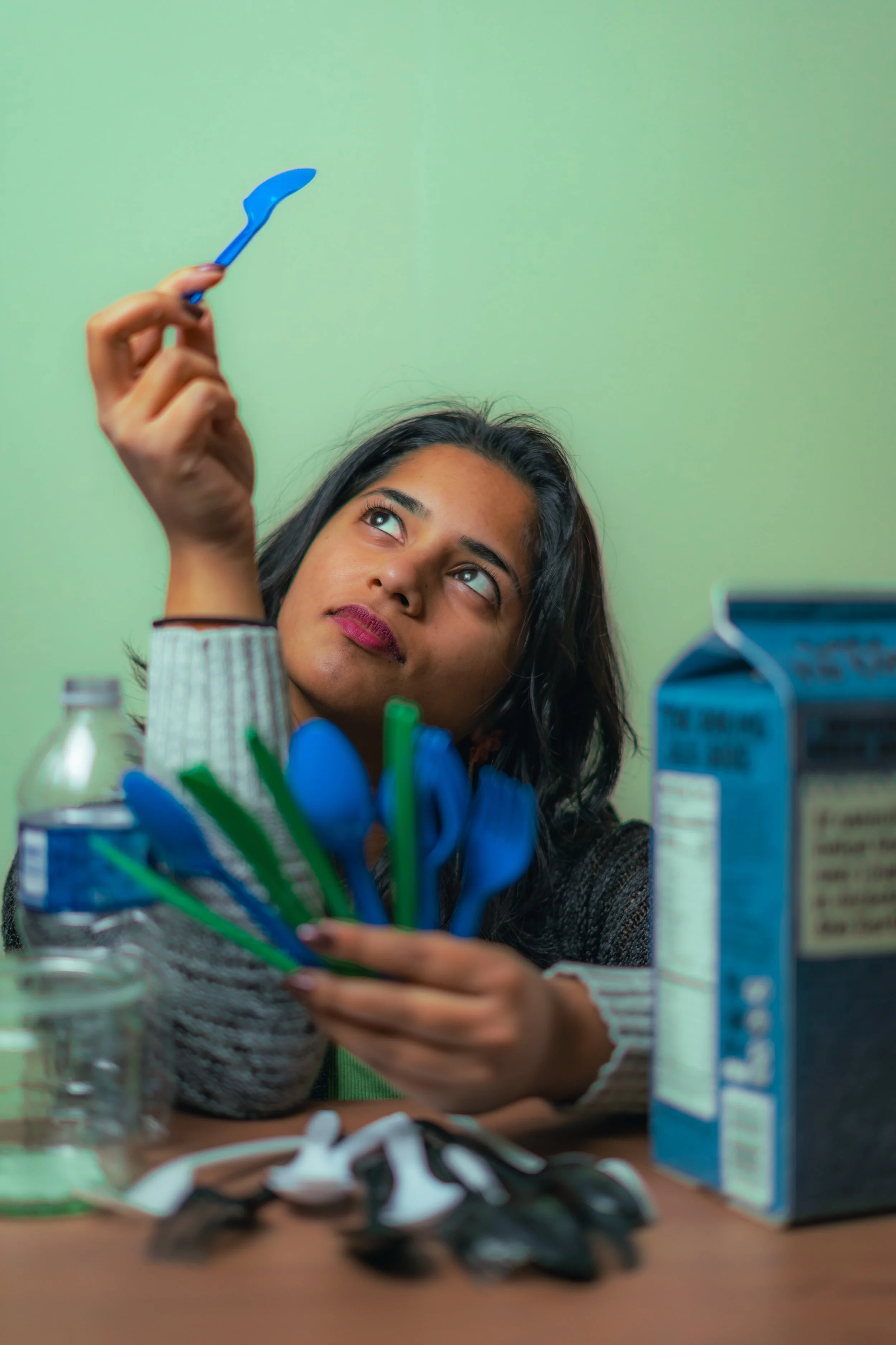 Young woman holding a blue spoon, looking up thoughtfully, with kitchen utensils on the table and a green wall background.