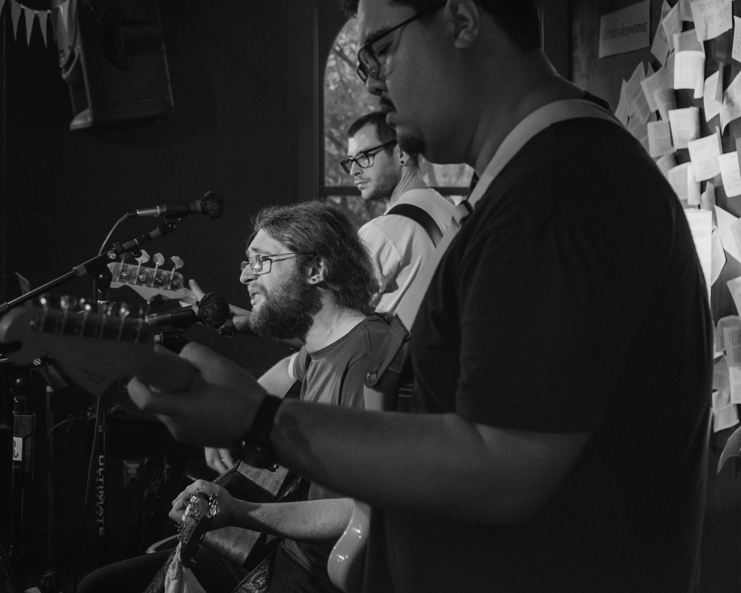 Four men are performing on stage with guitars and singing into microphones, with notes or papers on the wall behind them.