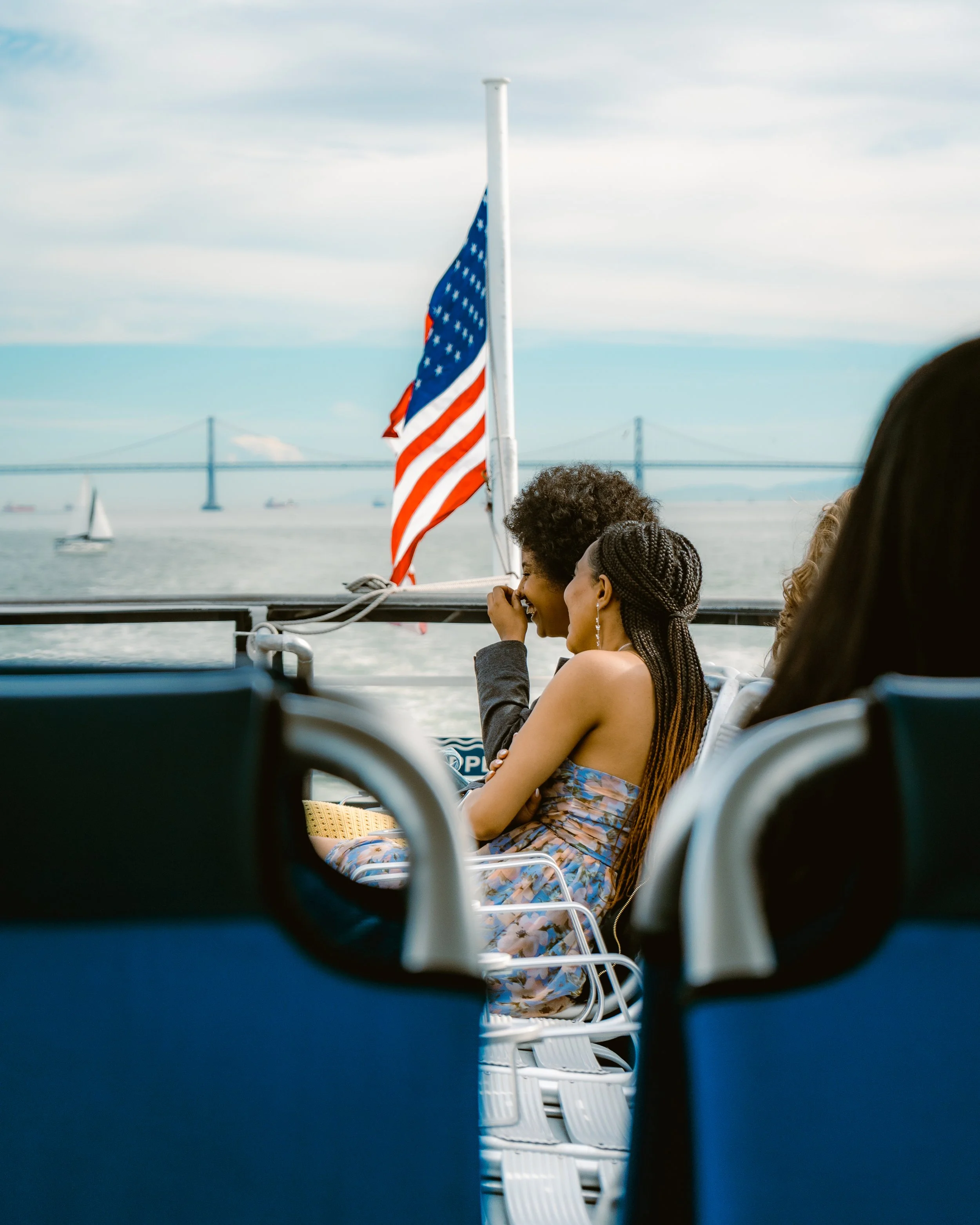 People sitting on a boat with an American flag, a bridge, and a sailboat on the water in the background.
