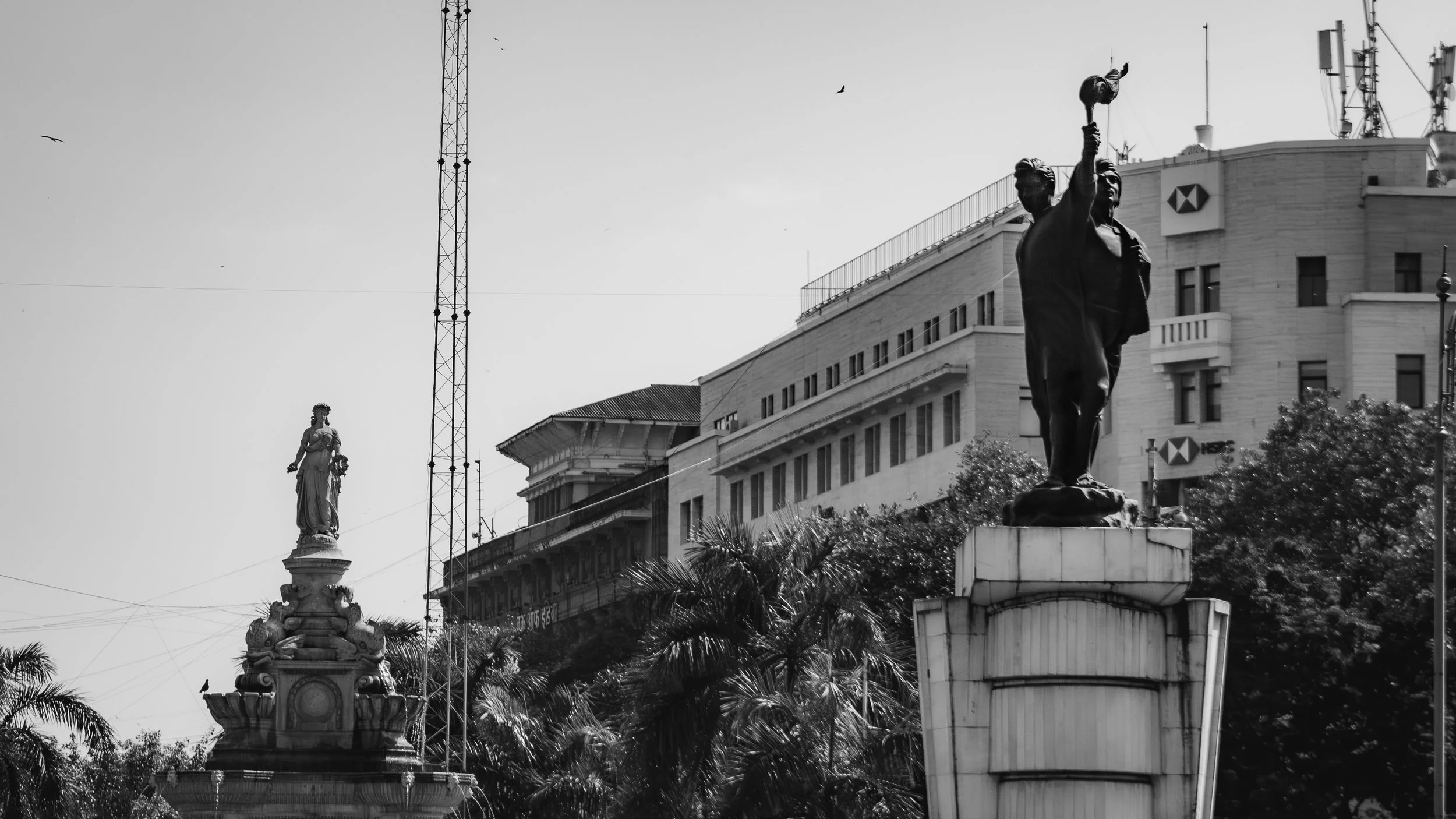 Black and white photo of two statues on tall pedestals in an urban setting, with trees and modern buildings in the background.
