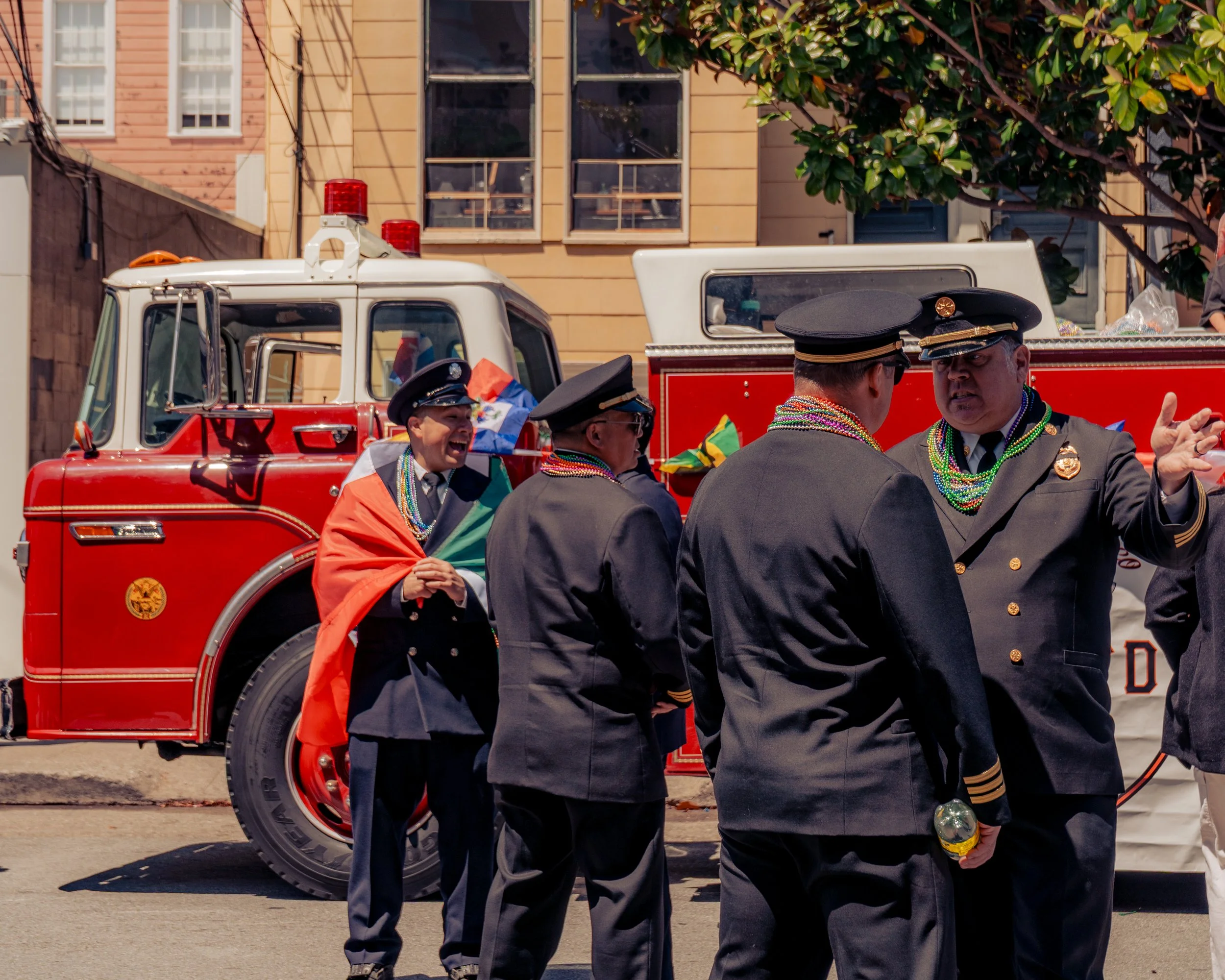 Group of uniformed men, likely firefighters, standing in front of a red fire truck during a celebration or parade. One man is draped in a rainbow-colored flag and wearing multiple beads, engaged in conversation with others.