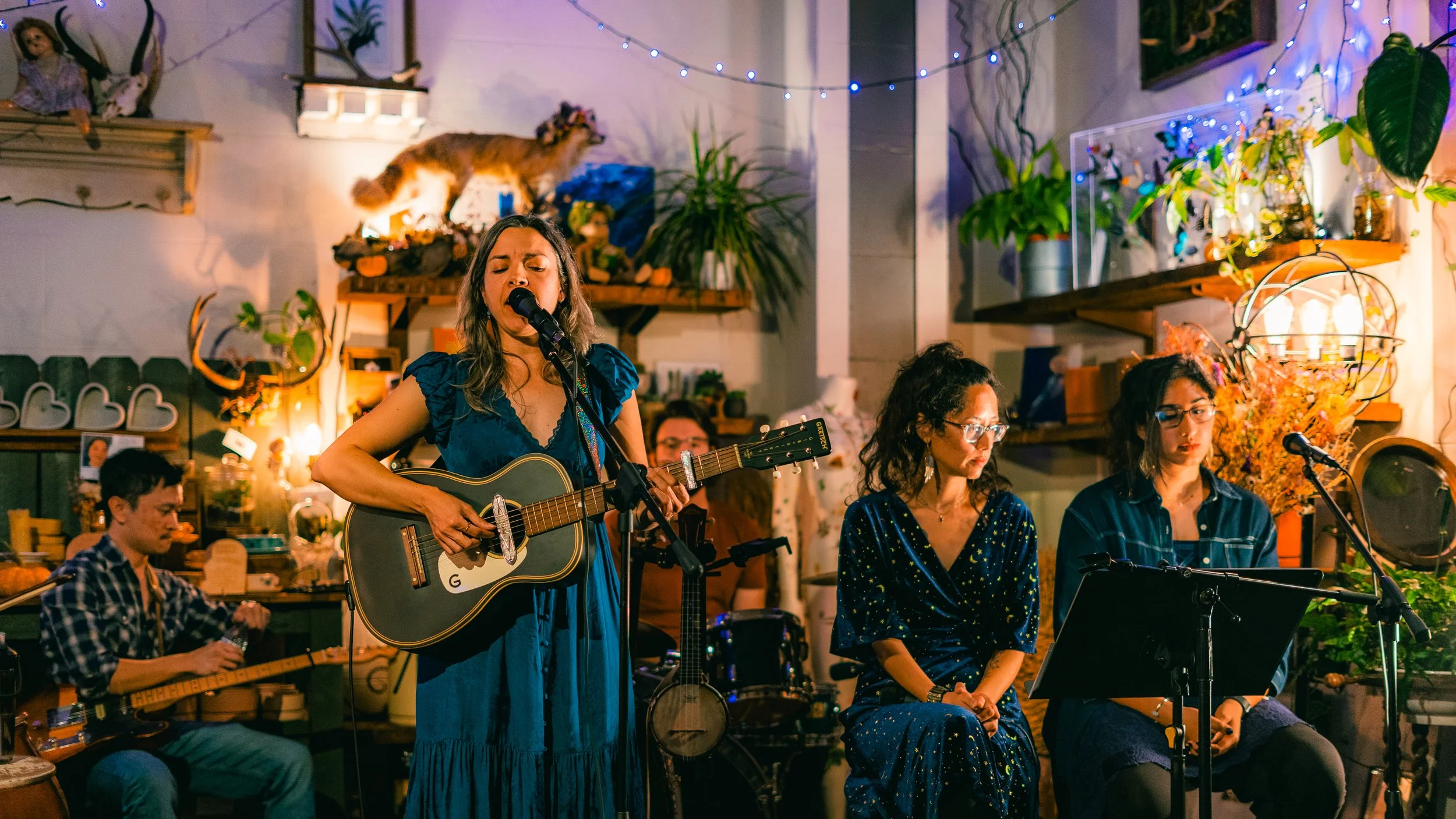 A woman with long hair in a blue dress is singing and playing a guitar, accompanied by three women sitting and a man playing a stringed instrument in a cozy, decorated indoor setting with plants and fairy lights.