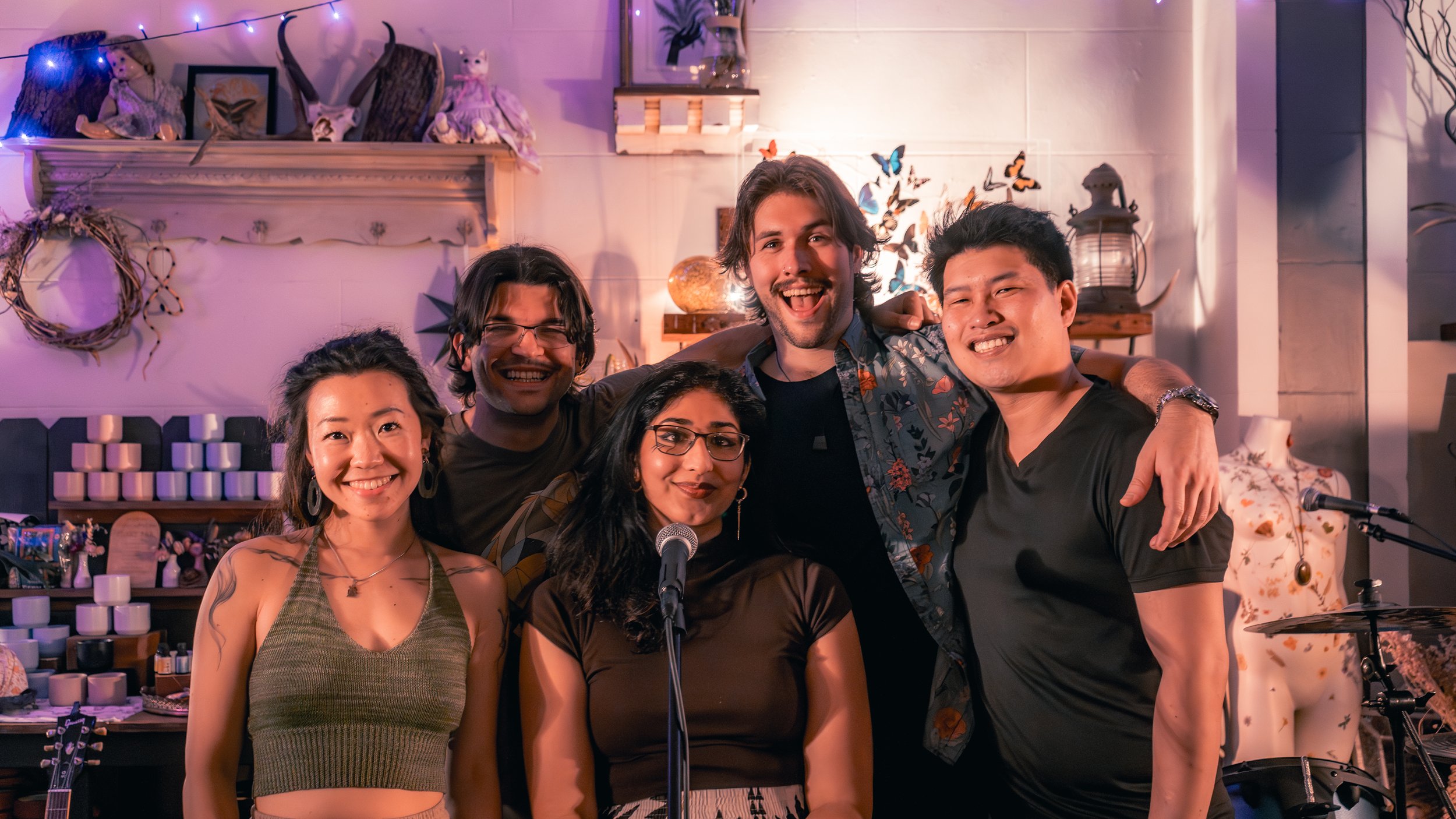 Group of six people smiling and posing for a photo indoors, with musical instruments, decorative items, and colorful butterfly wall art in the background.