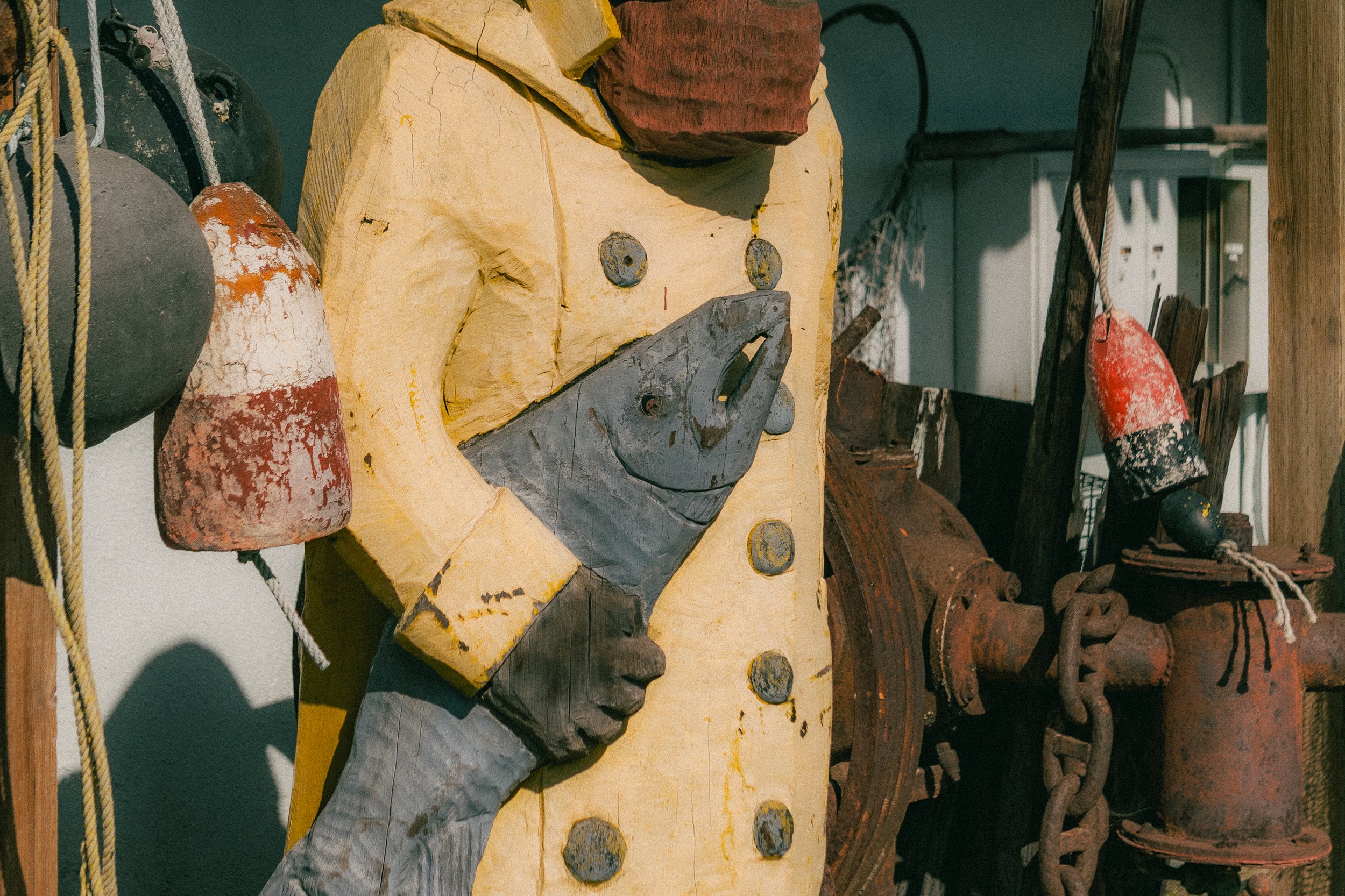 A colorful sculpture of a character with yellow, gray, and red painted parts, holding a fish-shaped object in its hand, surrounded by rusty industrial objects and ropes.