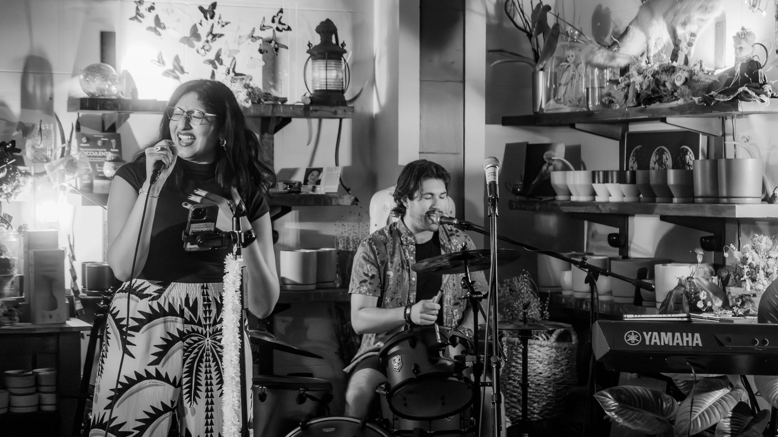 A woman singing into a microphone and a man playing the electronic drum set in a cozy, decorated room with shelves filled with plants, books, and decorative items.
