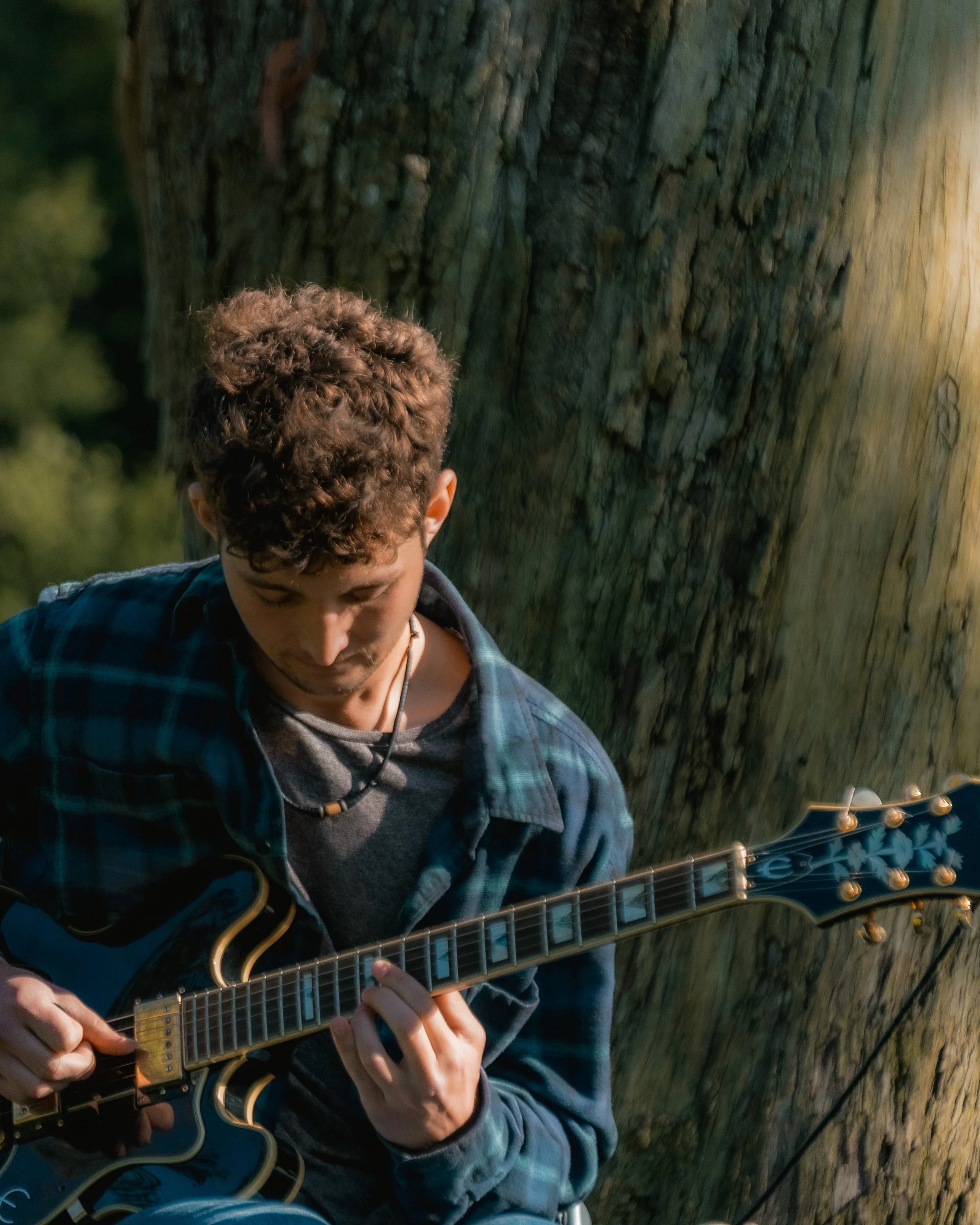 A young man with curly hair playing an electric guitar outdoors next to a large tree.