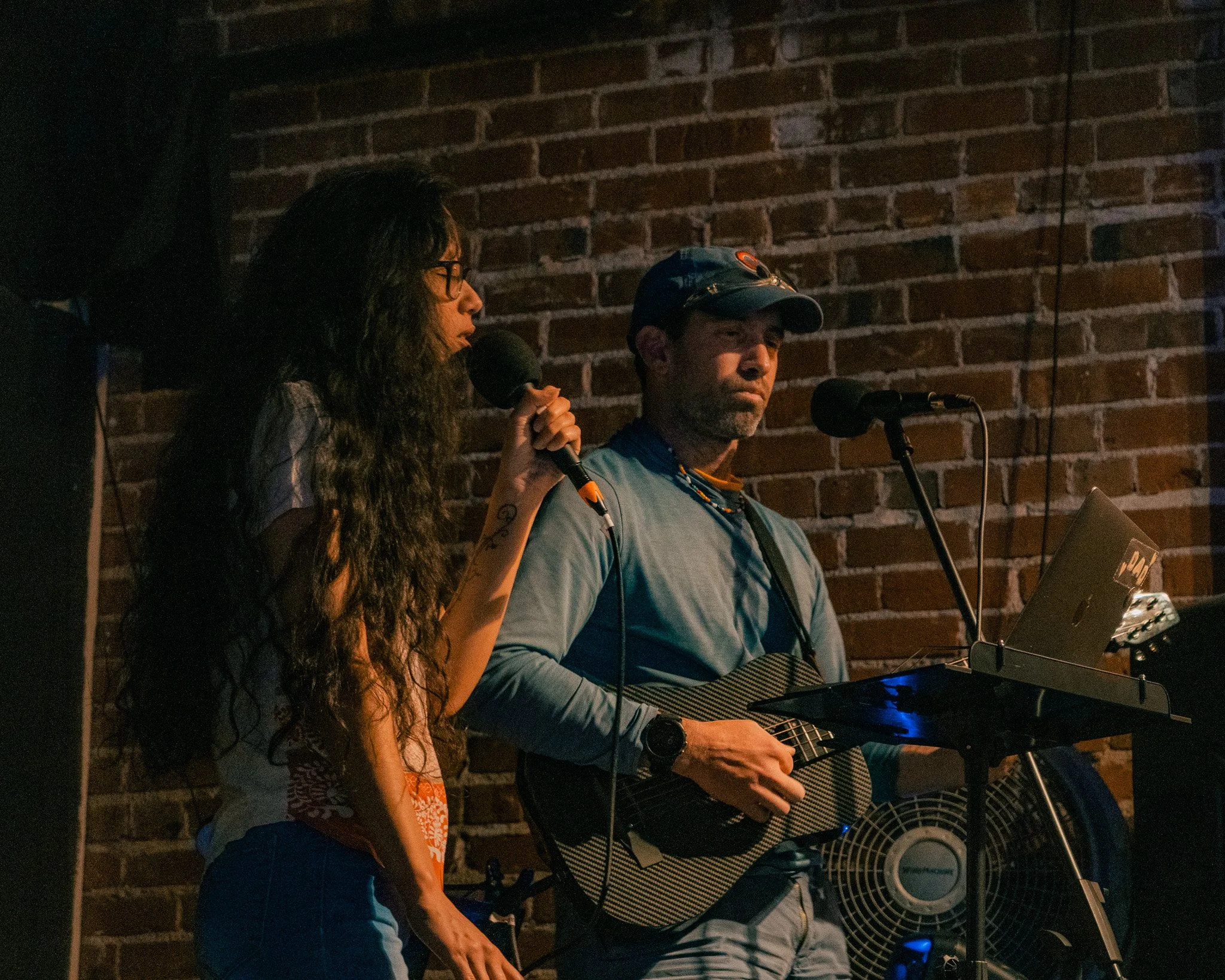 A woman with long wavy hair and glasses is singing into a microphone, and a man wearing a cap and a blue shirt is playing an acoustic guitar, both standing in front of a brick wall.