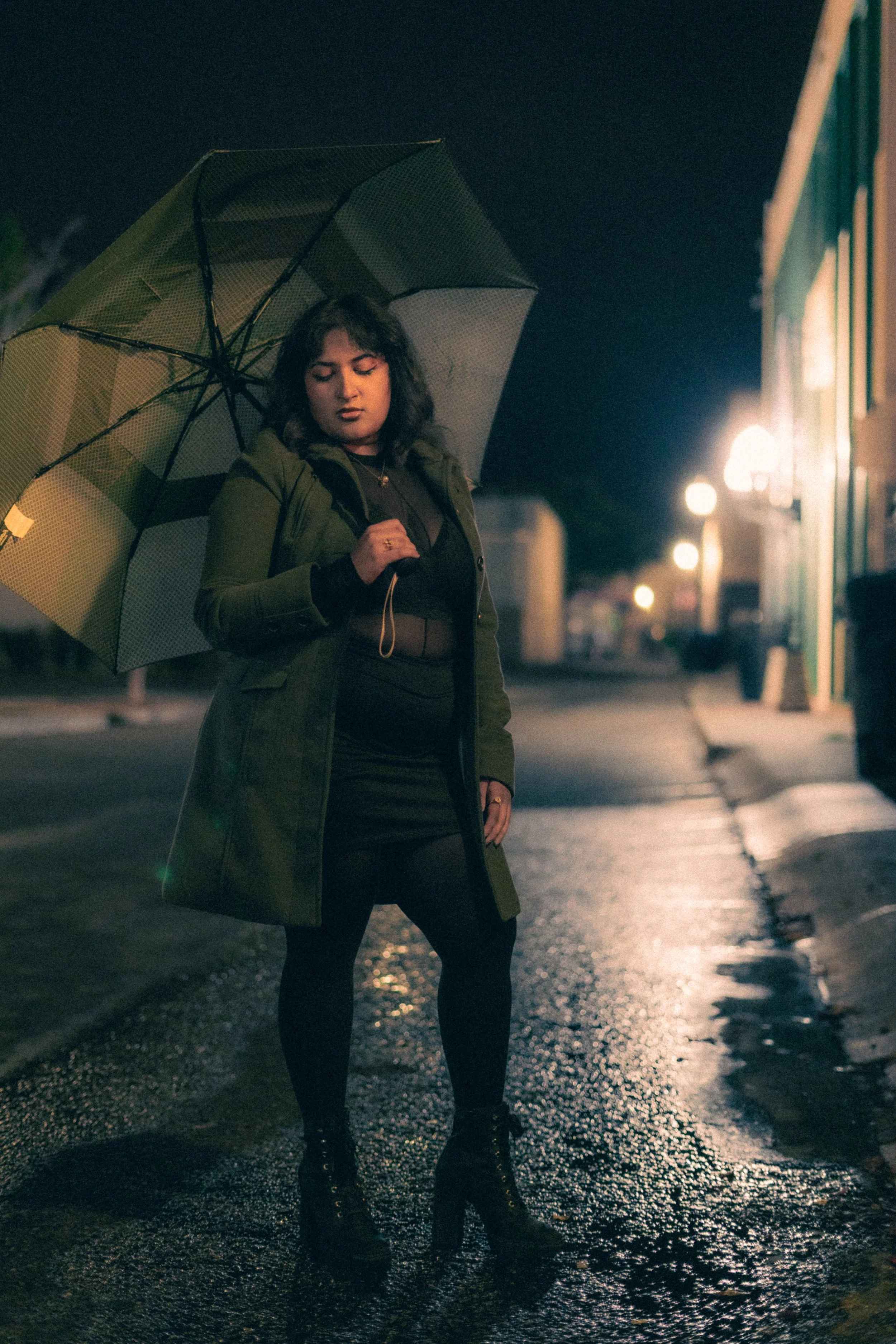 A woman standing on a wet street at night holding an umbrella, wearing a green coat, black dress, and black high boots.