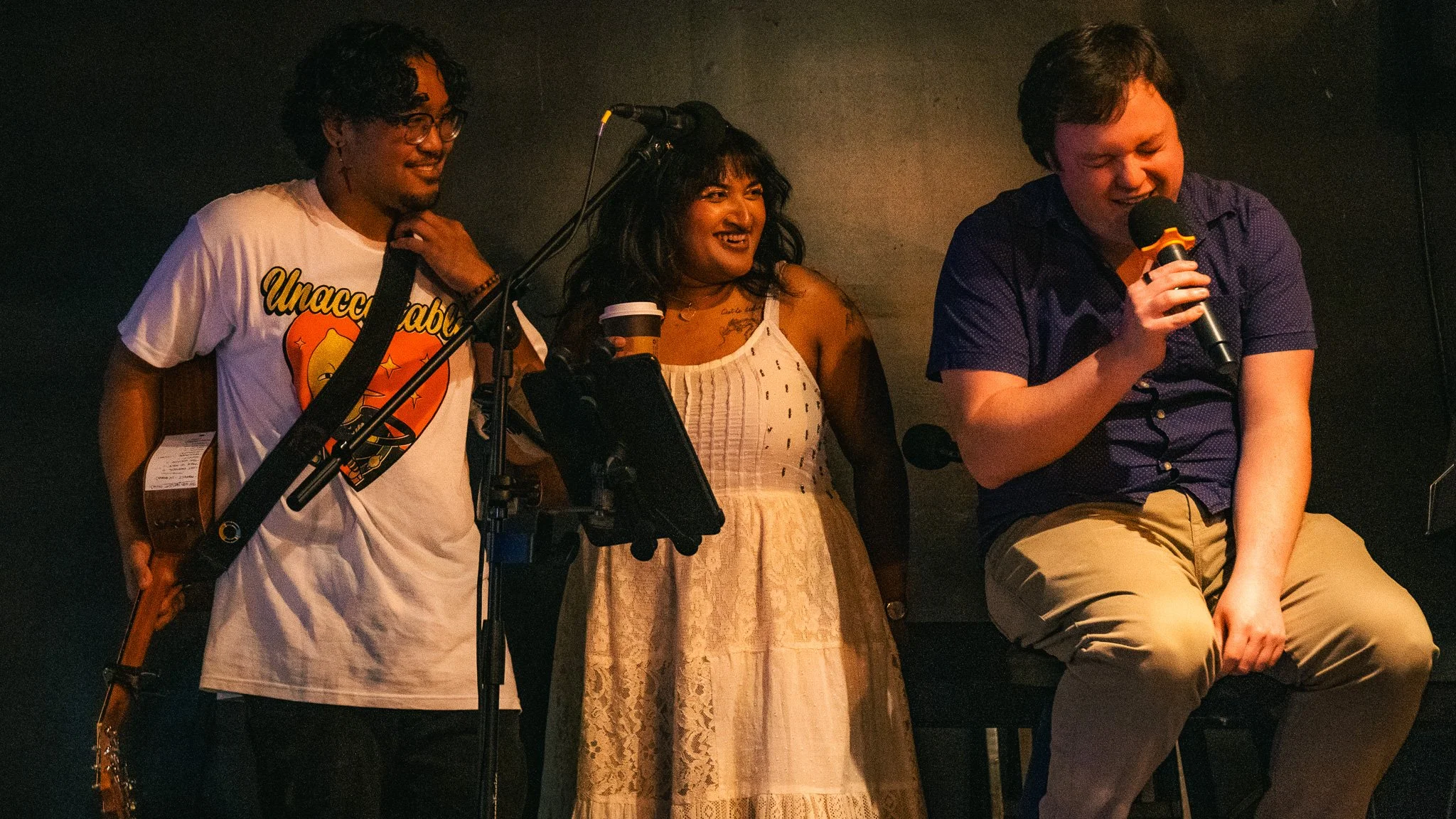 Three people on a stage, two men and one woman, appear in a performance setting with microphones and musical instruments. The woman is smiling and holding a coffee cup, while one man is holding a microphone and appears to be singing or speaking, and 