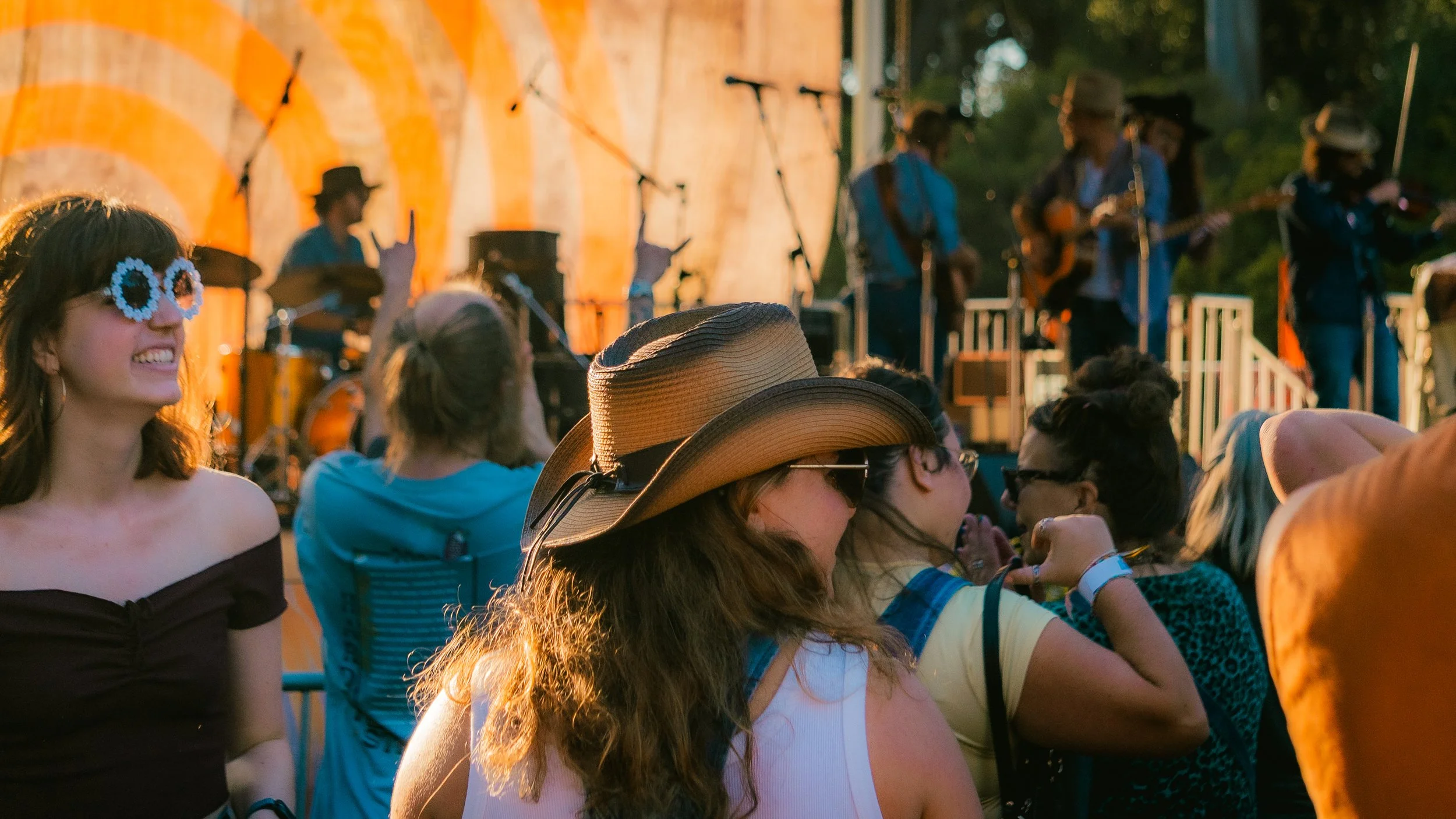 People attending an outdoor music concert, some wearing sunglasses and hats, enjoying the live band performing on stage.