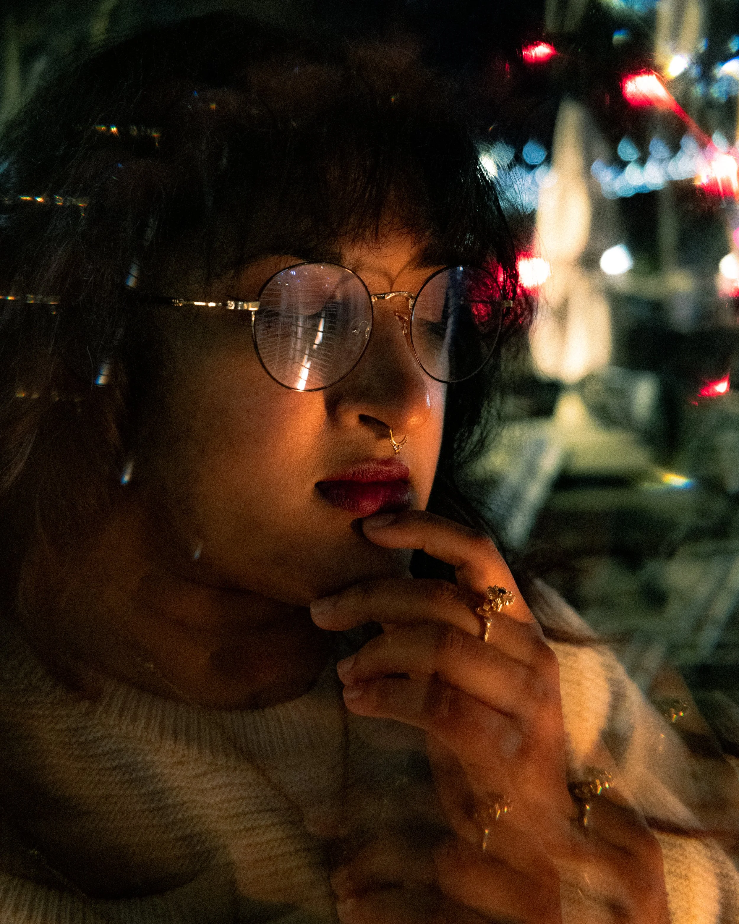Portrait of a woman with dark hair and glasses with round lenses, tilting her head slightly with her finger near her lips. She is wearing rings, a septum piercing, and a light-colored sweater, with a blurred city street background at night with light