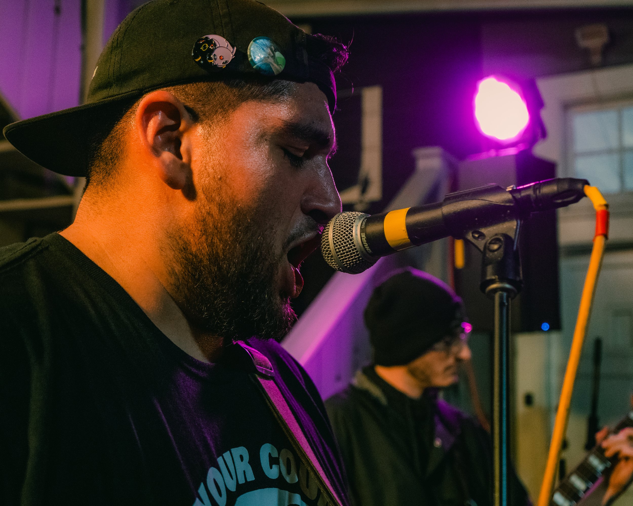A male musician with a beard, wearing a black cap with pins, sings into a microphone during a performance with purple stage lighting.