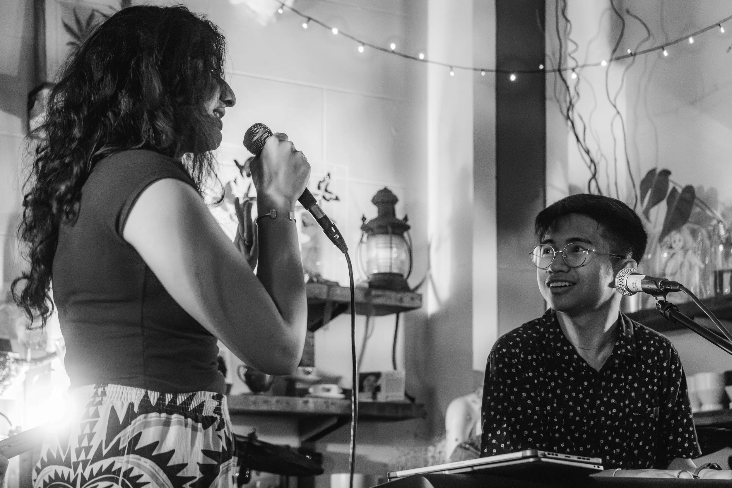 A woman and a man are performing music together in a cozy, decorated indoor space. The woman, standing on the left, is singing into a microphone, while the man, sitting on the right, is playing a keyboard and looking at her. The background features s