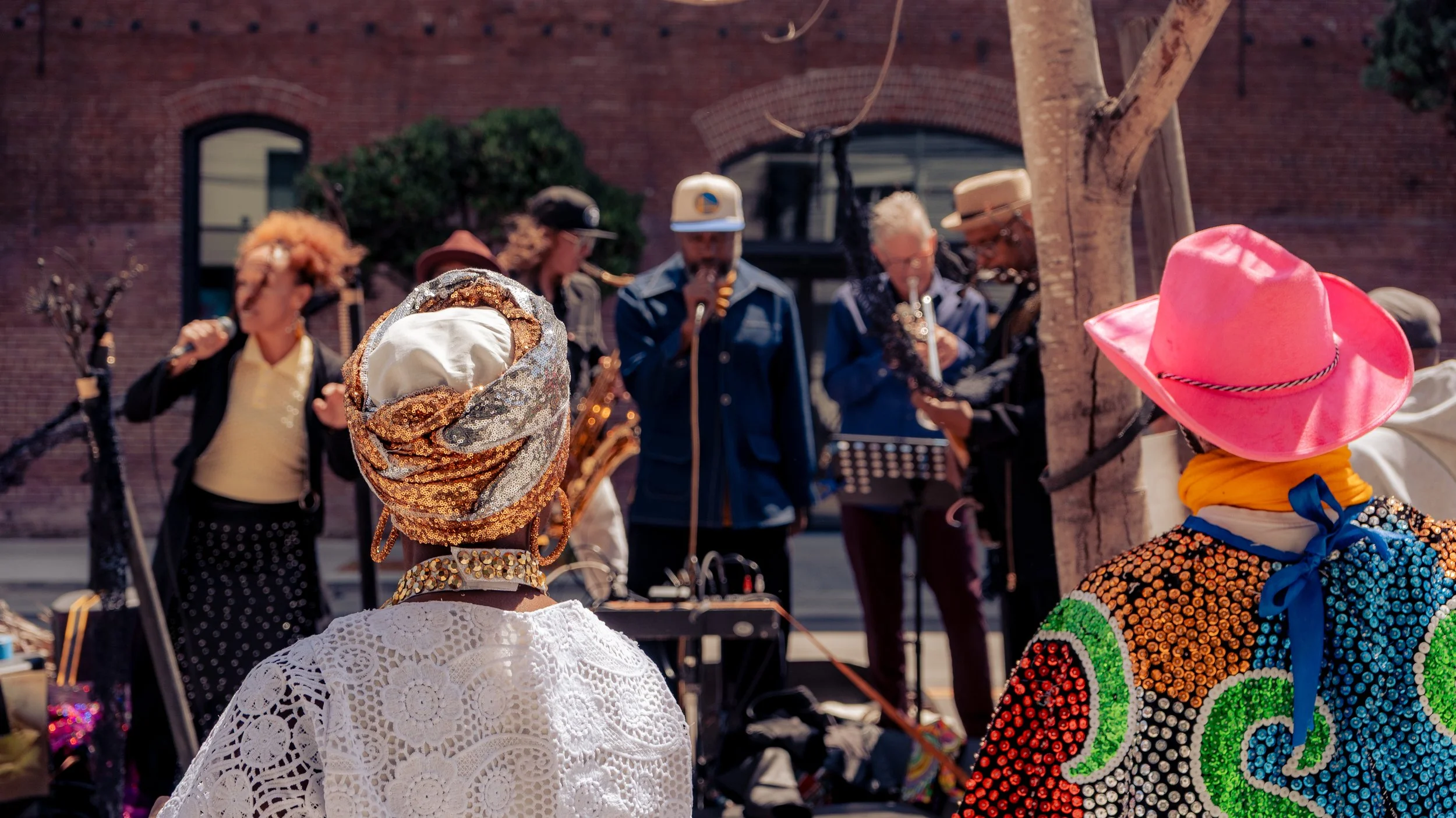 People performing music outdoors, with two women in colorful hats and one with a lace outfit in the foreground and a band playing in the background.