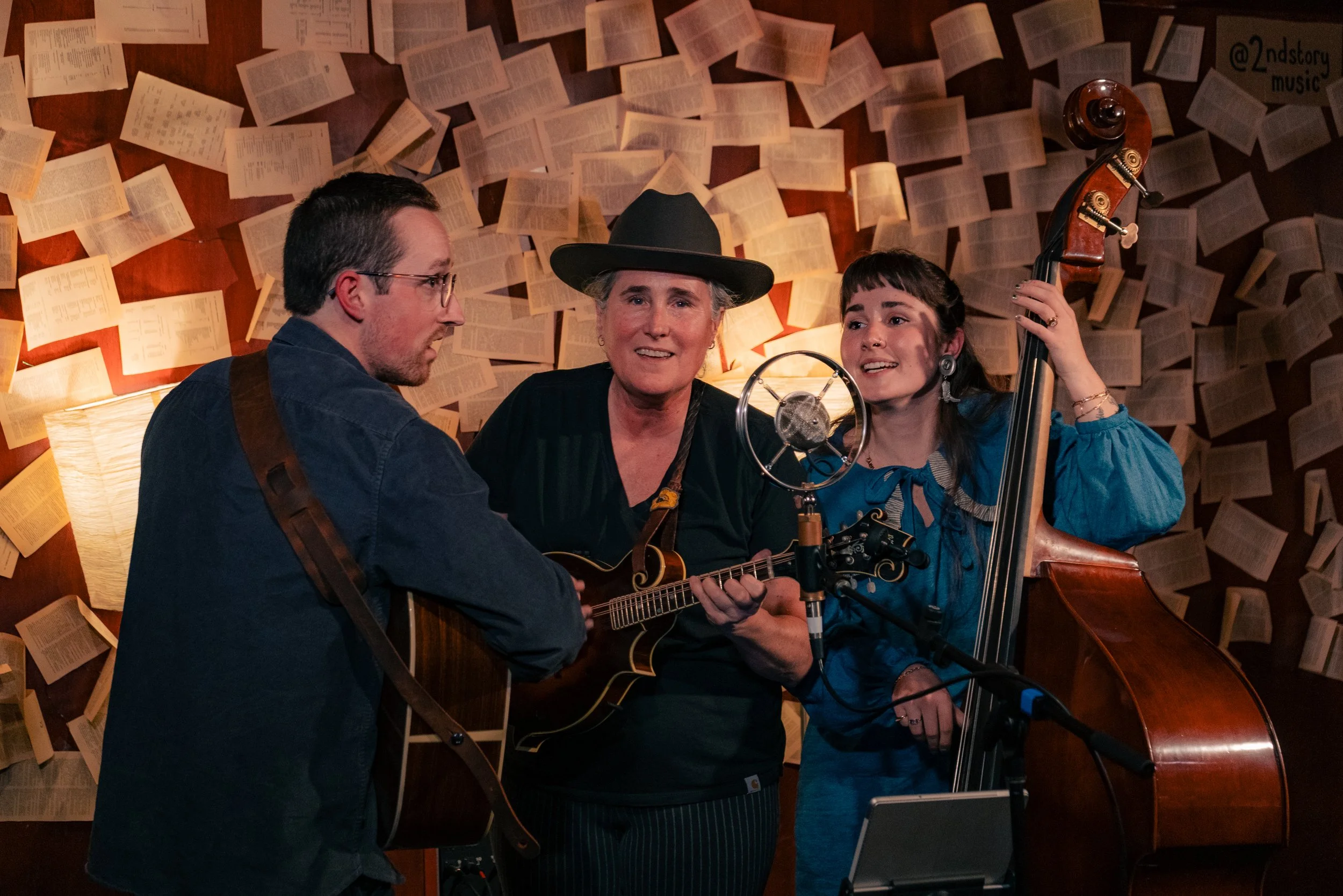 Three musicians performing in a cozy room with books on the wall, a man playing guitar, a woman with a mandolin, and a woman with a double bass, singing and engaging with each other.