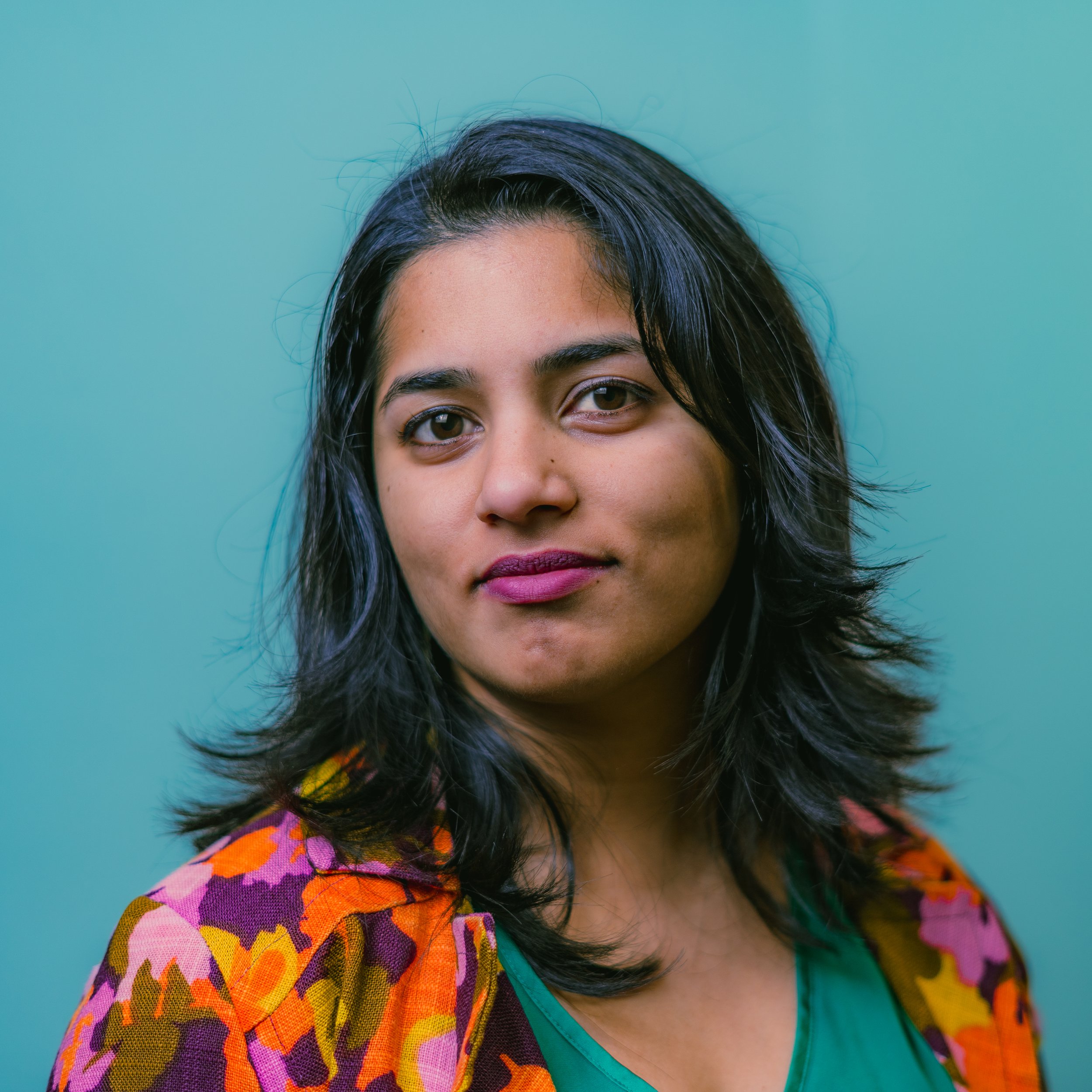 Portrait of a woman with medium skin tone, dark wavy hair, pink lipstick, wearing a green top and a colorful floral blazer.