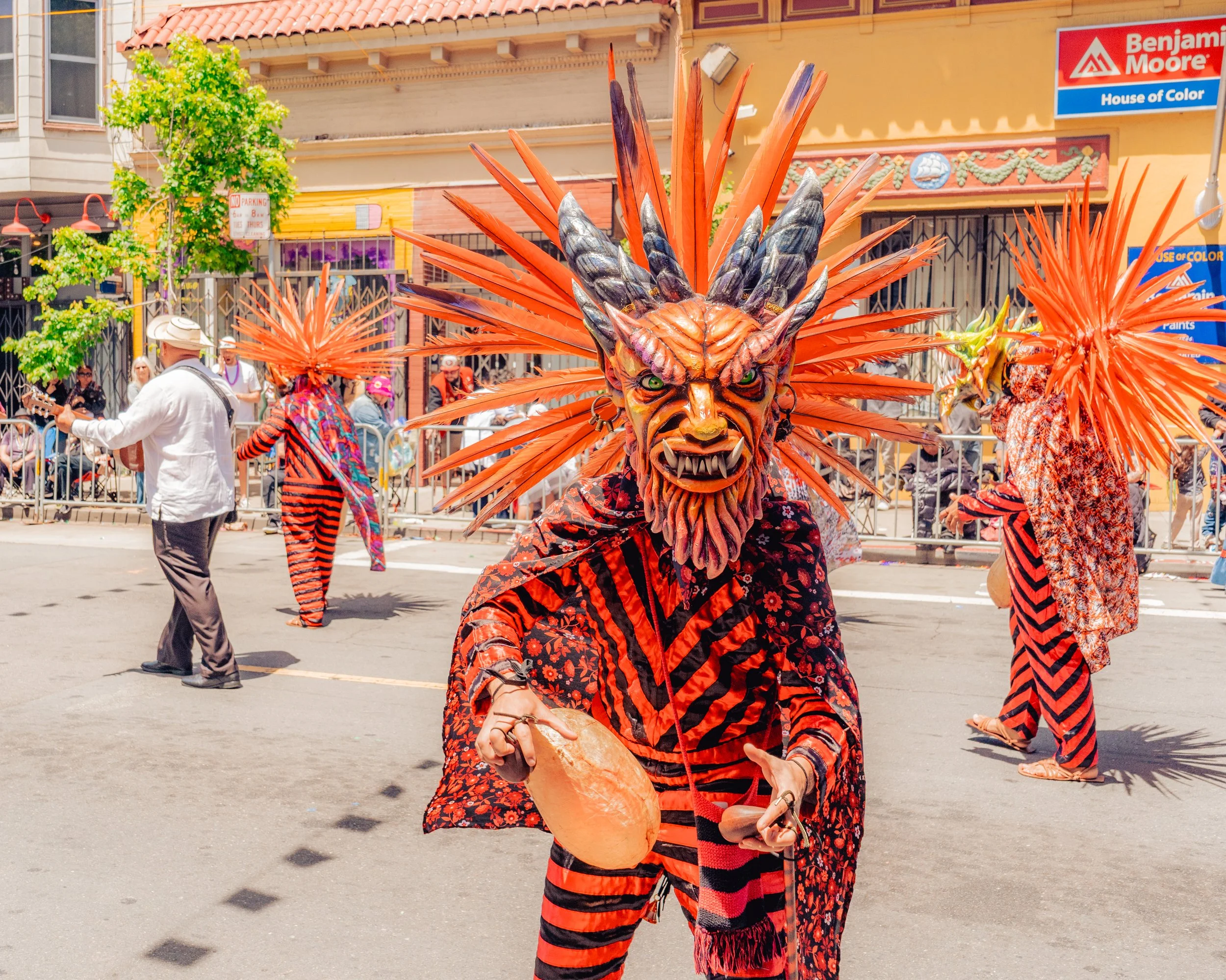 Person dressed in a colorful, elaborate costume with a fierce, demonic mask and large orange feathered headdress participating in a street parade.