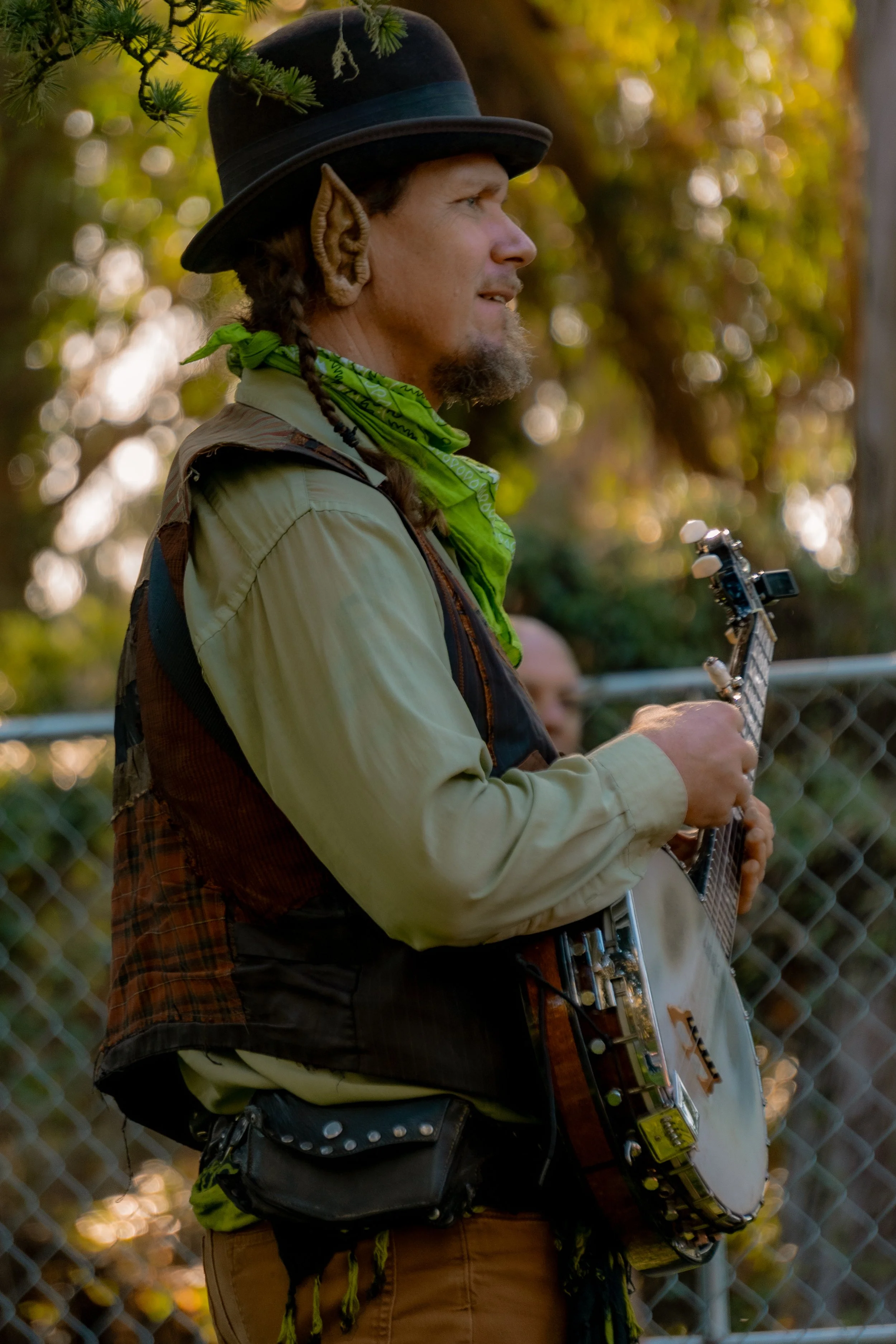 A man with elf ears playing a guitar outdoors during daytime, with a chain-link fence and trees in the background.