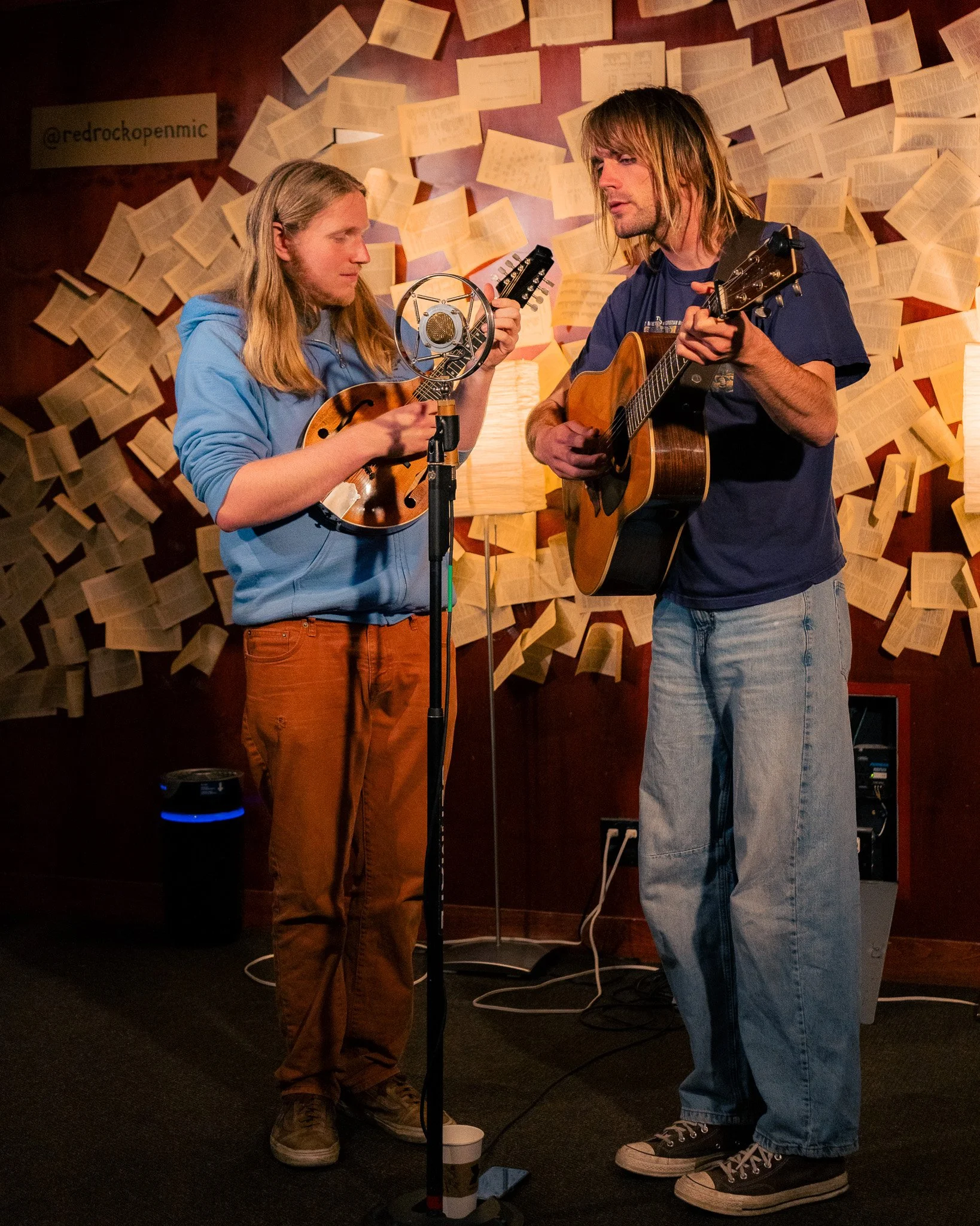 Two men are playing guitars and singing into a microphone in a cozy room decorated with open books on the wall.