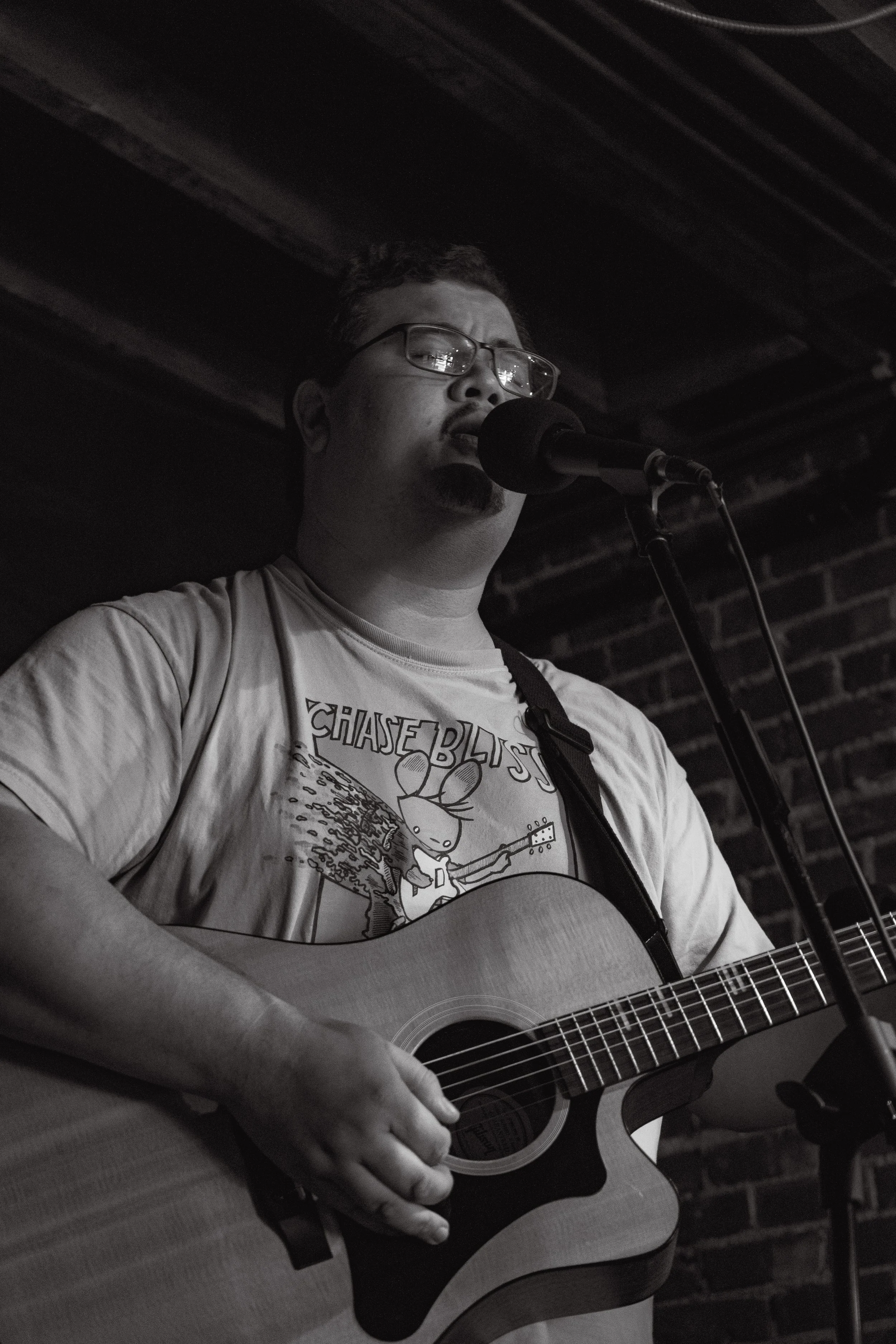 A man wearing glasses and a Chase Blast T-shirt playing an acoustic guitar and singing into a microphone in a dimly lit venue with a brick wall background.