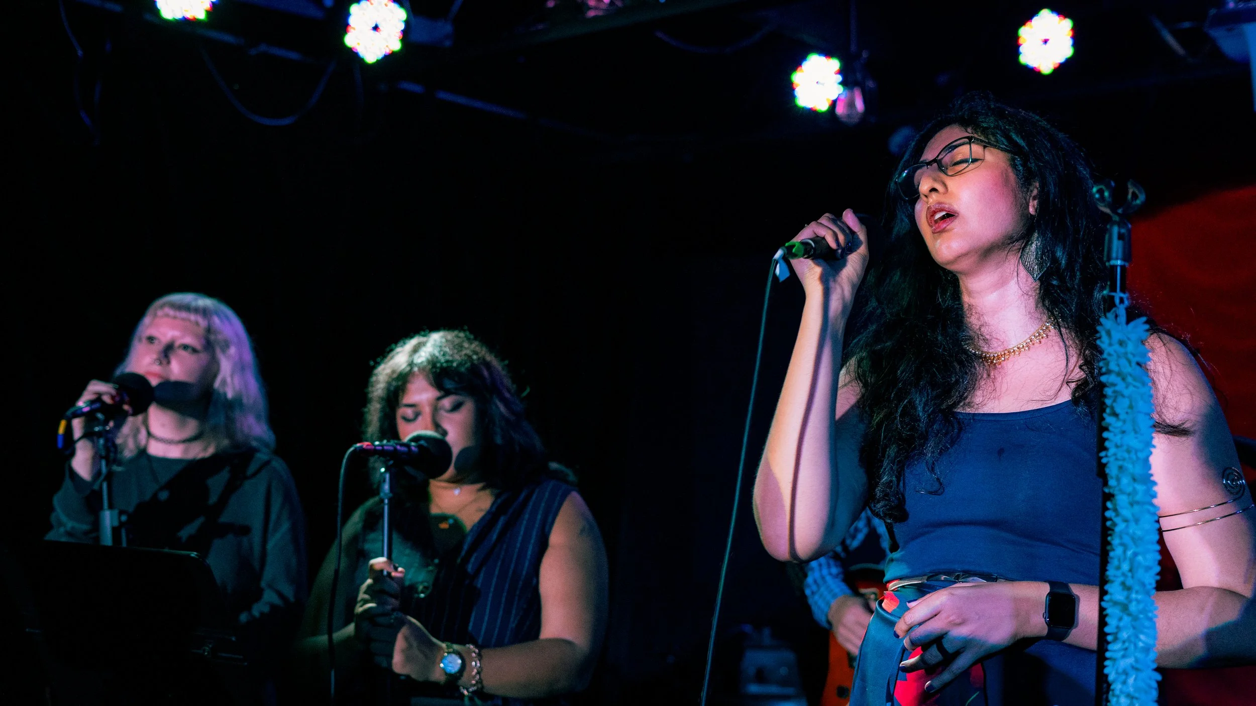 Three women singing into microphones on a stage with colorful lights overhead.