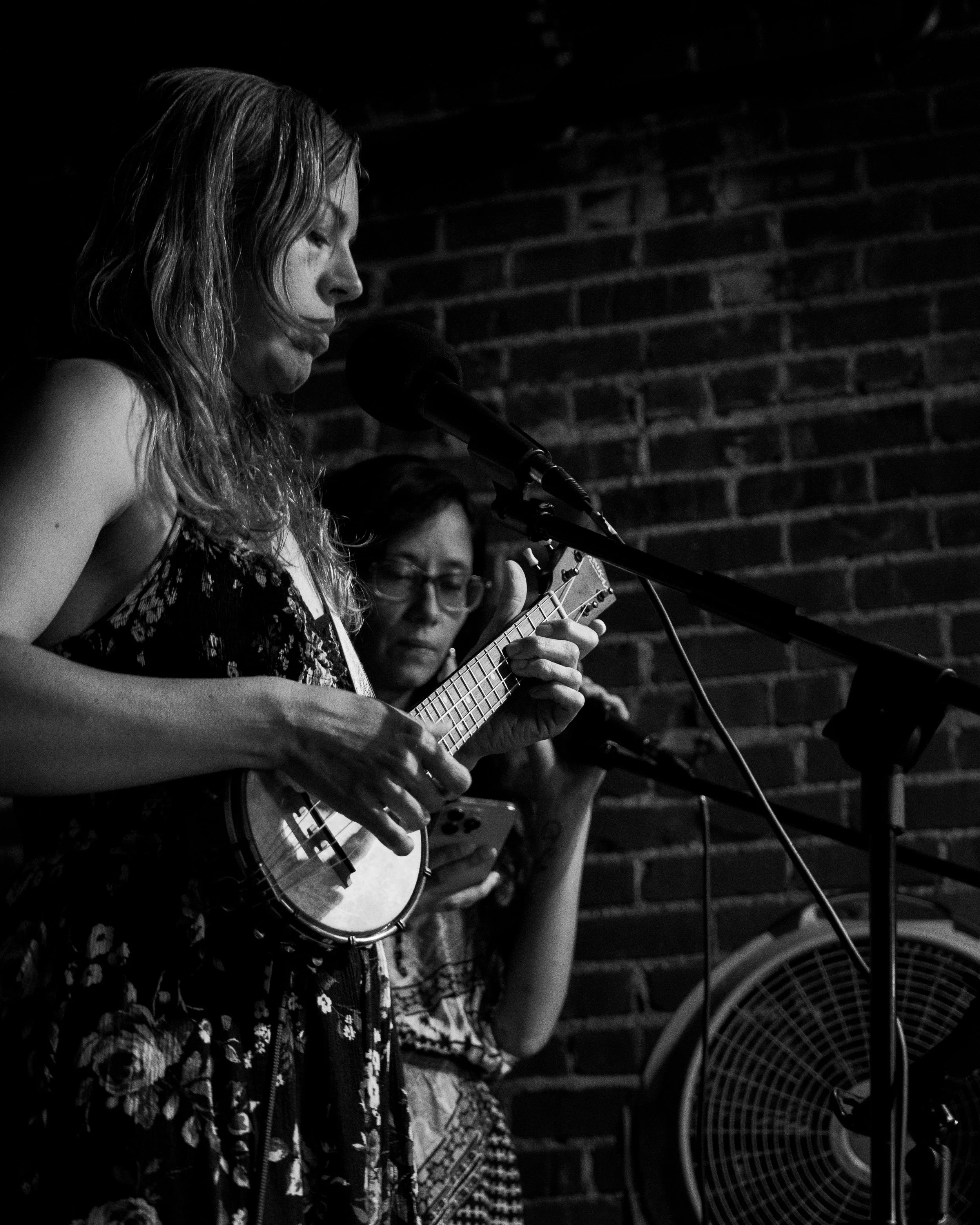 Two women performing on stage, one singing into a microphone and playing a ukulele, the other playing a viola and holding a phone, with a brick wall in the background.