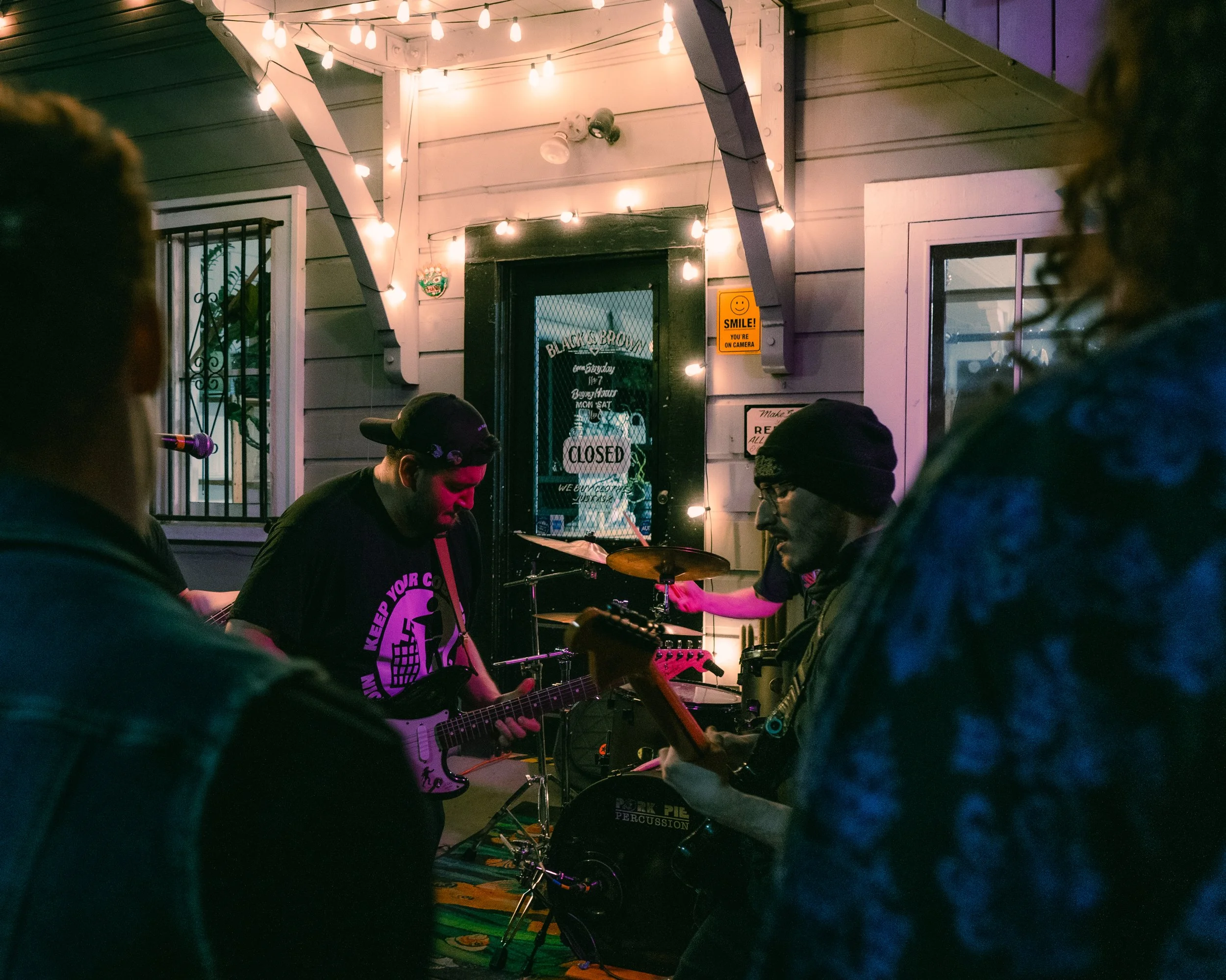 A small band performs outside at night, with string lights overhead. The band members are playing guitars and drums, and are gathered in front of a building with a sign that reads 'CLOSED'. There are several audience members watching the performance.