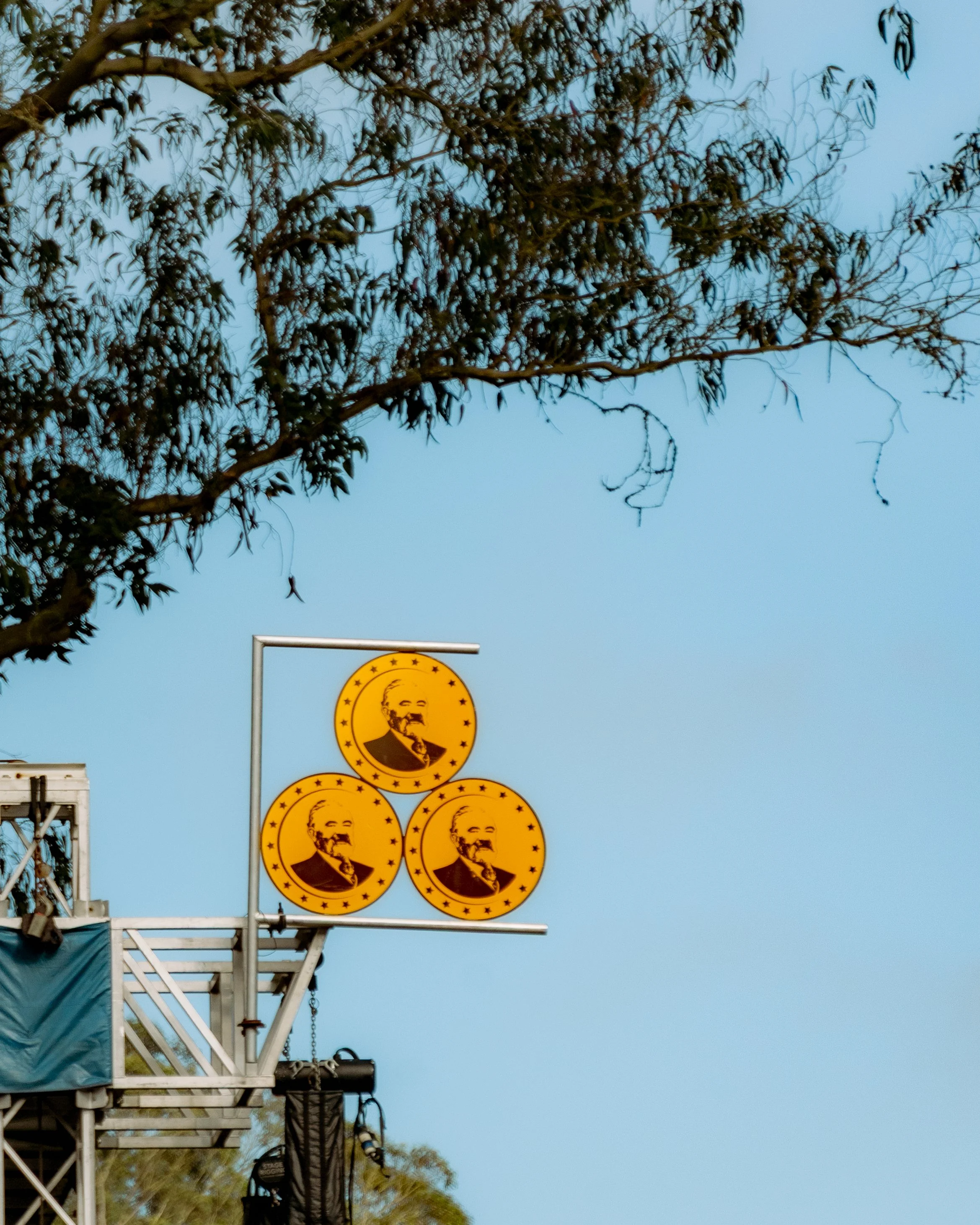 Three yellow circular signs with a black illustration of Karl Marx's profile, bordered with stars, mounted on a metal pole against a light blue sky, with tree branches above.