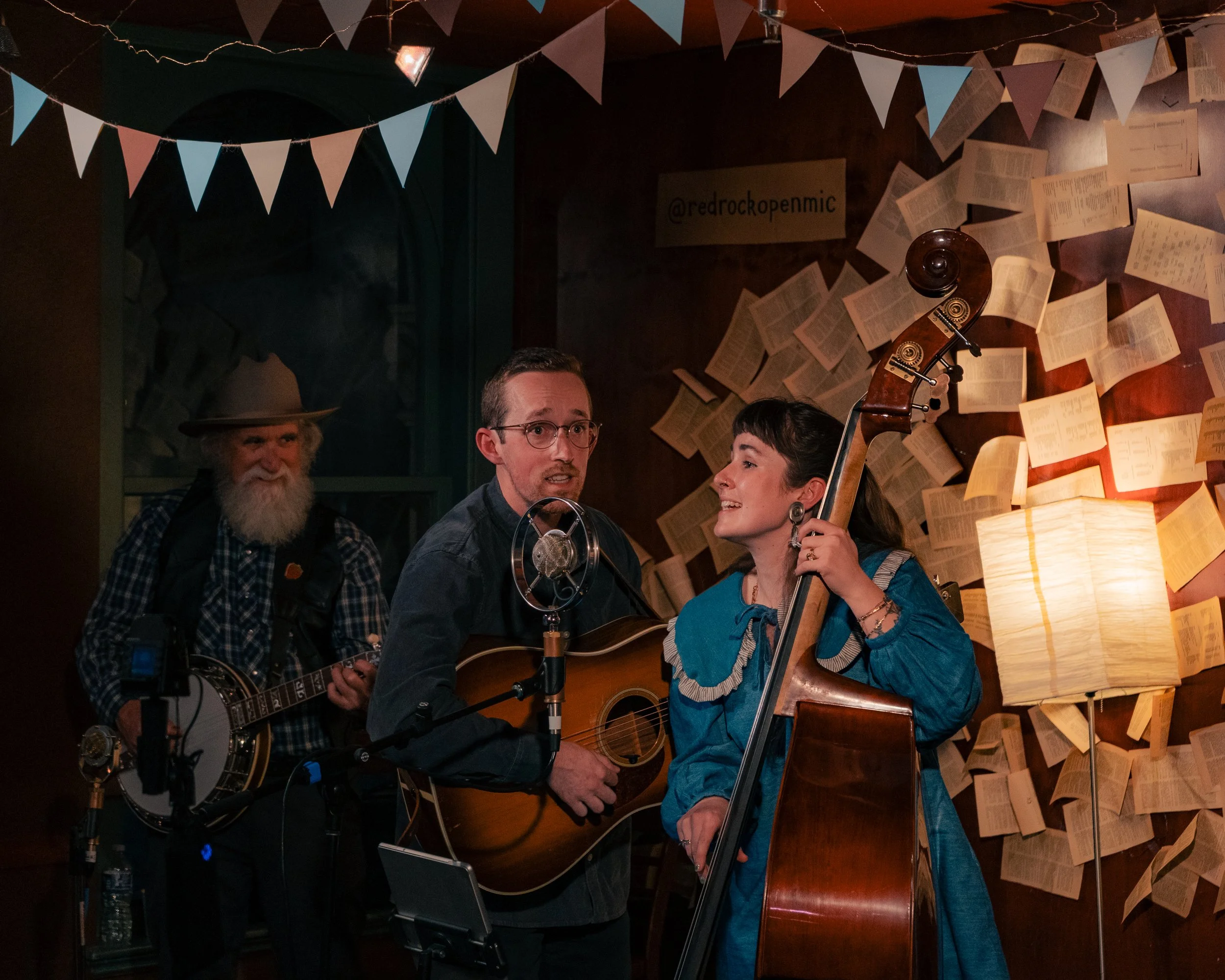 Three musicians performing indoors, with two men playing acoustic guitars and a woman playing upright bass. The background features open books on the wall, a lit lamp, and bunting decorations.