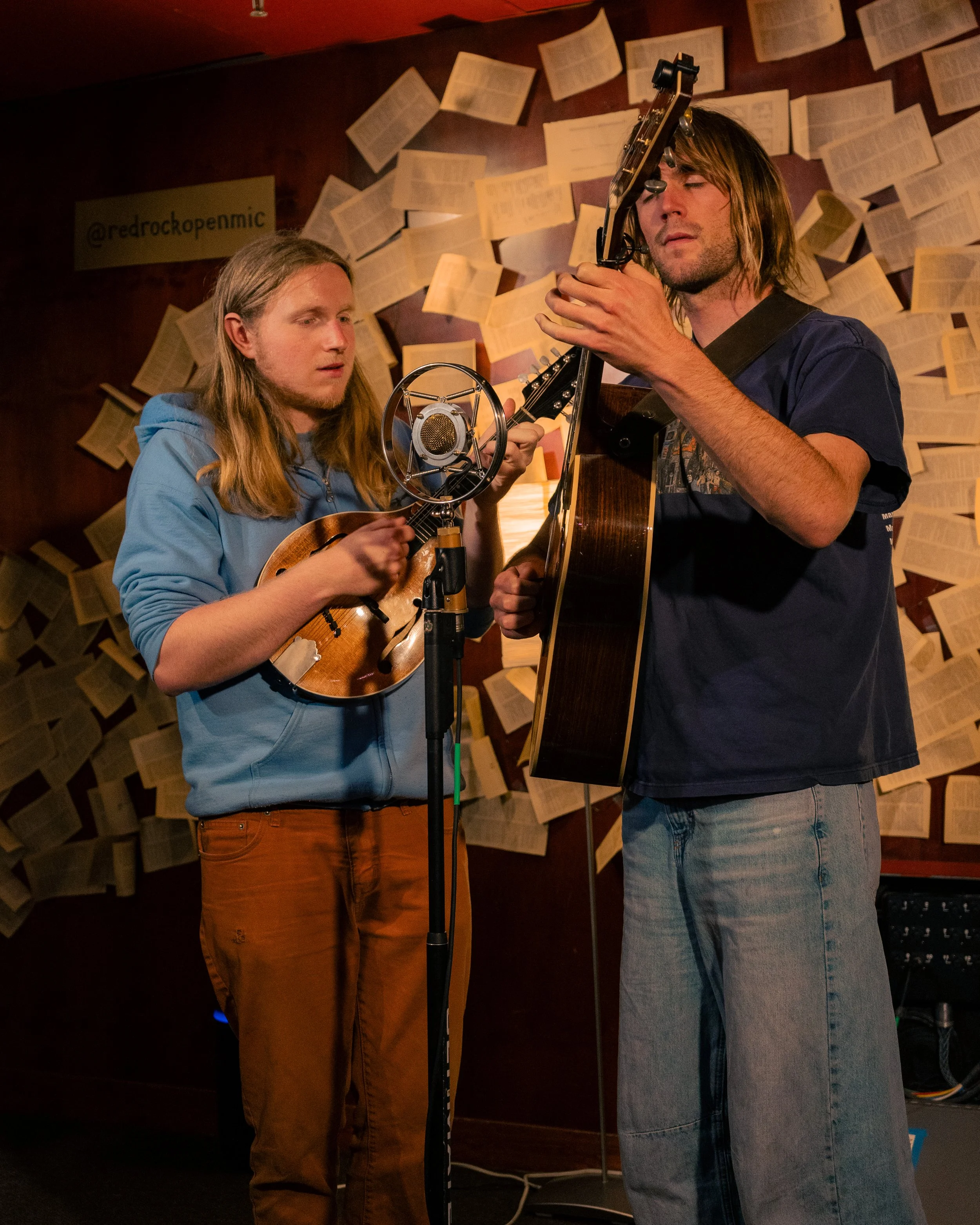 Two young men playing musical instruments, one with a mandolin and the other with an acoustic guitar, in a cozy room decorated with open books on the wall.