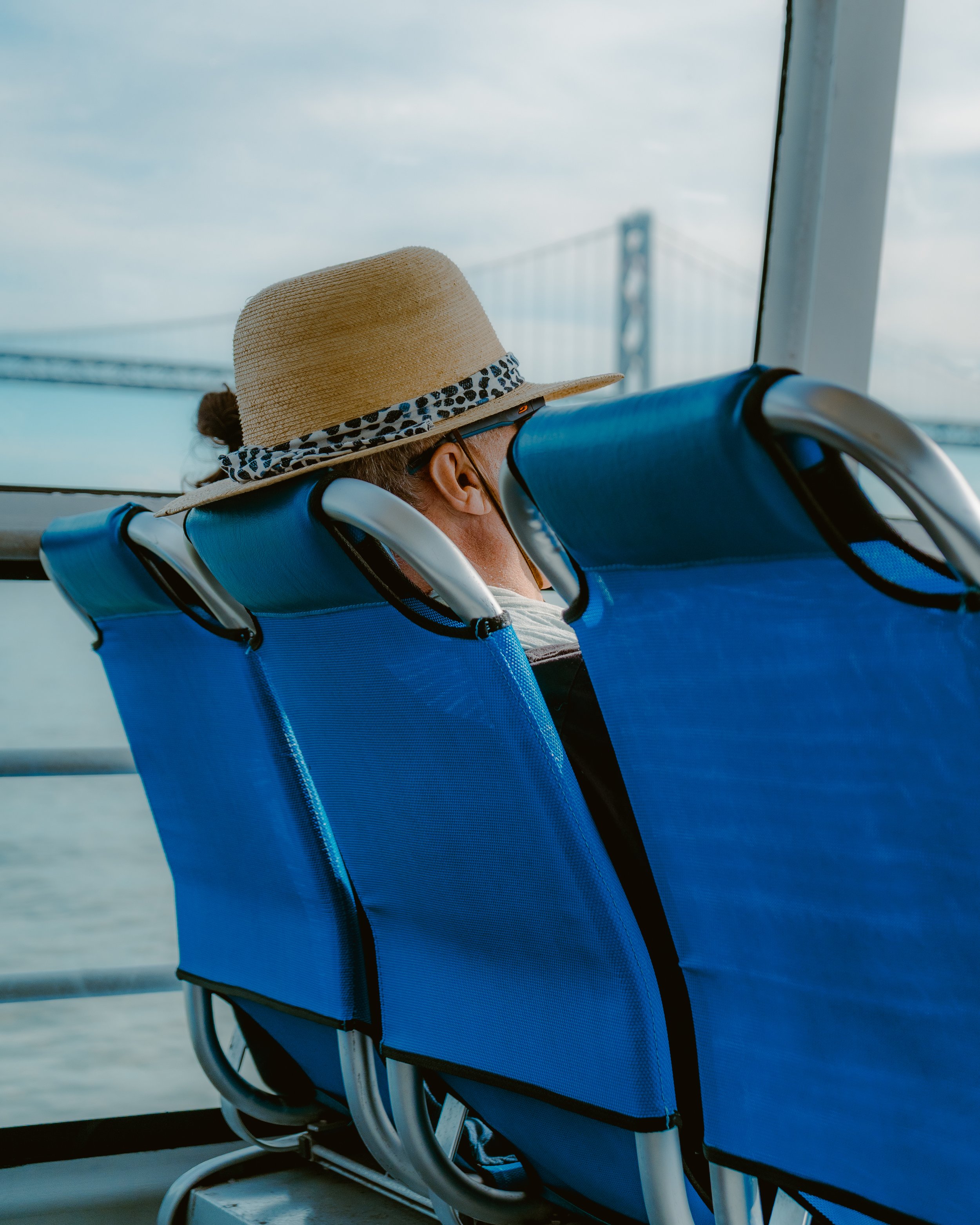 Person wearing a straw hat with a leopard print band, sitting on a blue seat on a ferry, with a view of a bridge and water outside the window.