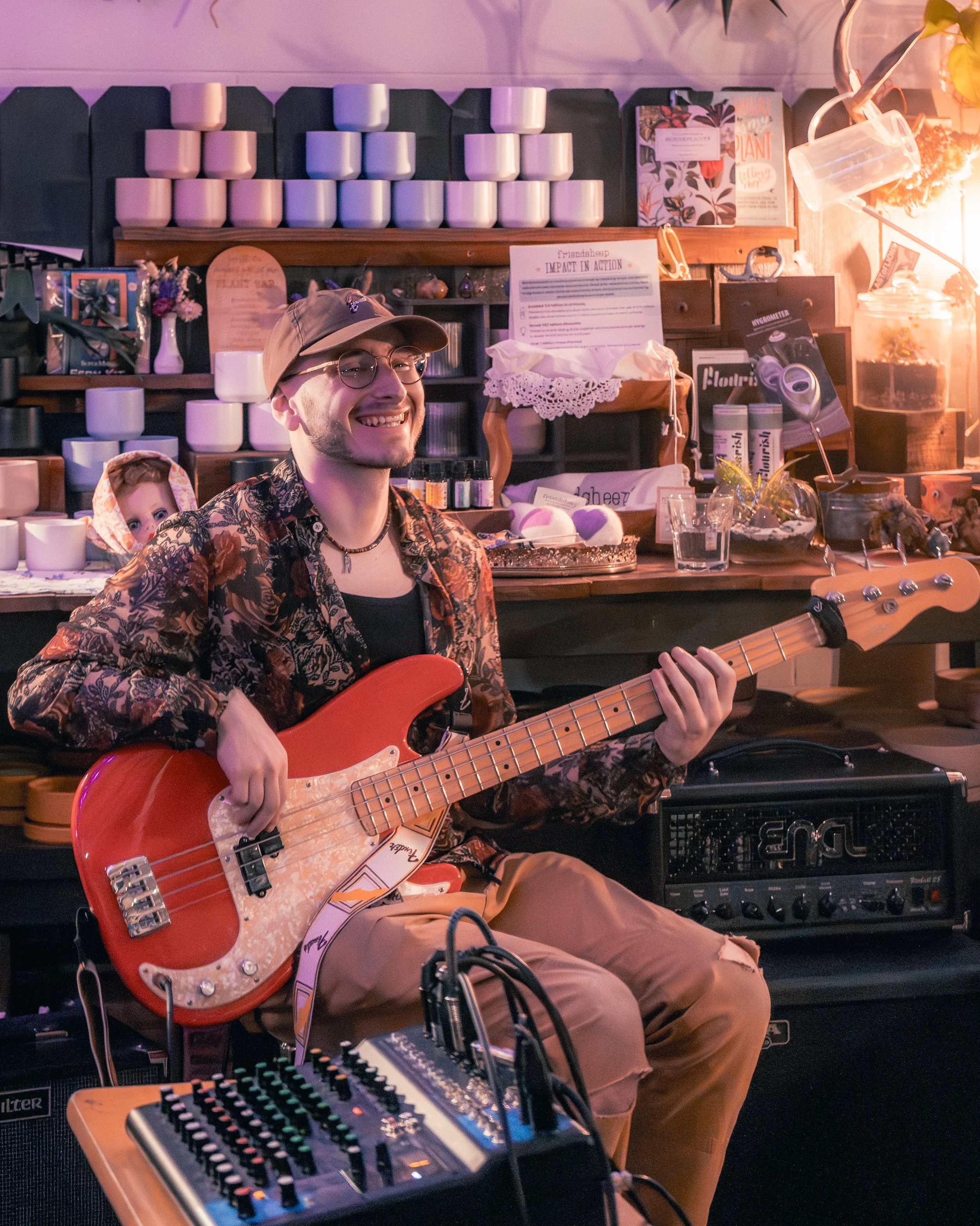 A smiling young man wearing glasses, a beige cap, and a floral shirt plays a red electric bass guitar. He sits in a cozy space with various decorative items on shelves behind him, including candles, books, and plants, and several music equipment piec