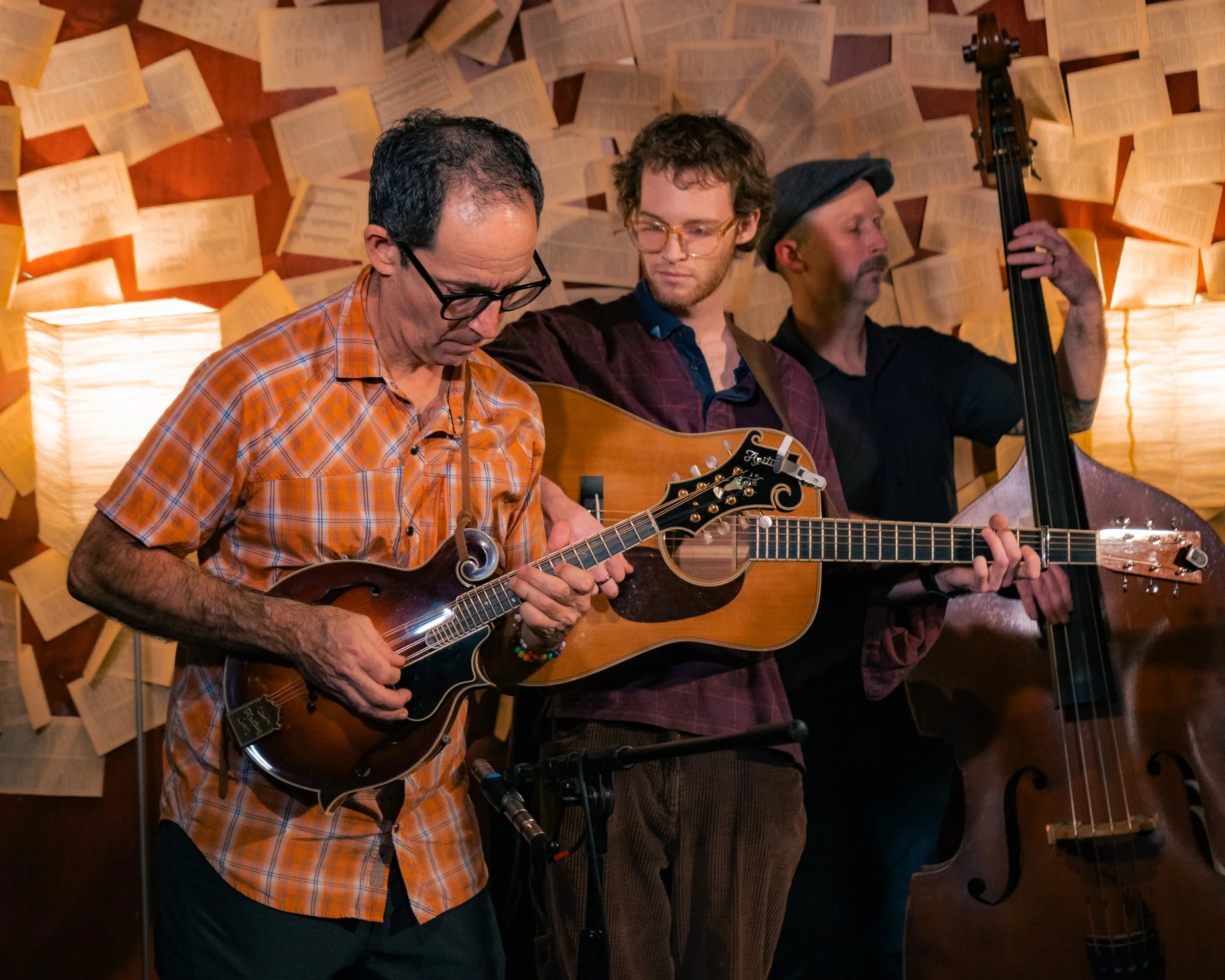 Three musicians playing string instruments, with sheet music or song lyrics on the wall behind them.