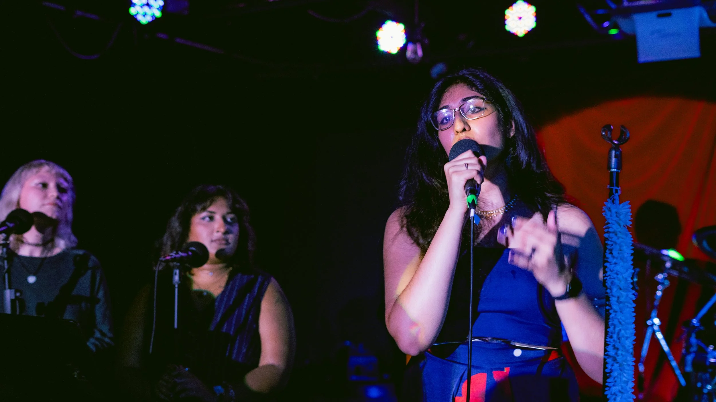 Three women singing on stage in a dimly lit venue with colorful lights overhead. The woman in front has long dark hair, glasses, and is holding a microphone close to her mouth.