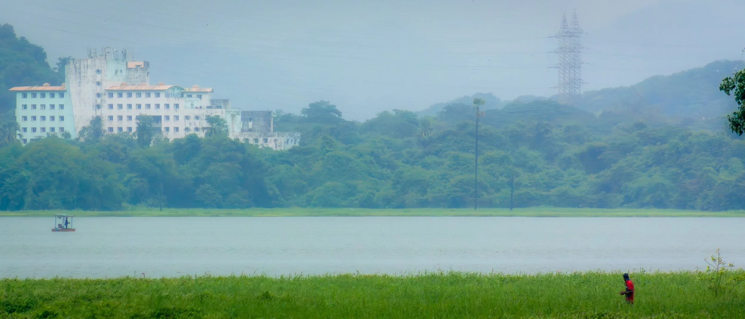 A person in a red shirt standing near a green grassy area by a lake, with a boat on the water, lush green trees, and buildings in the background behind a hazy atmosphere.