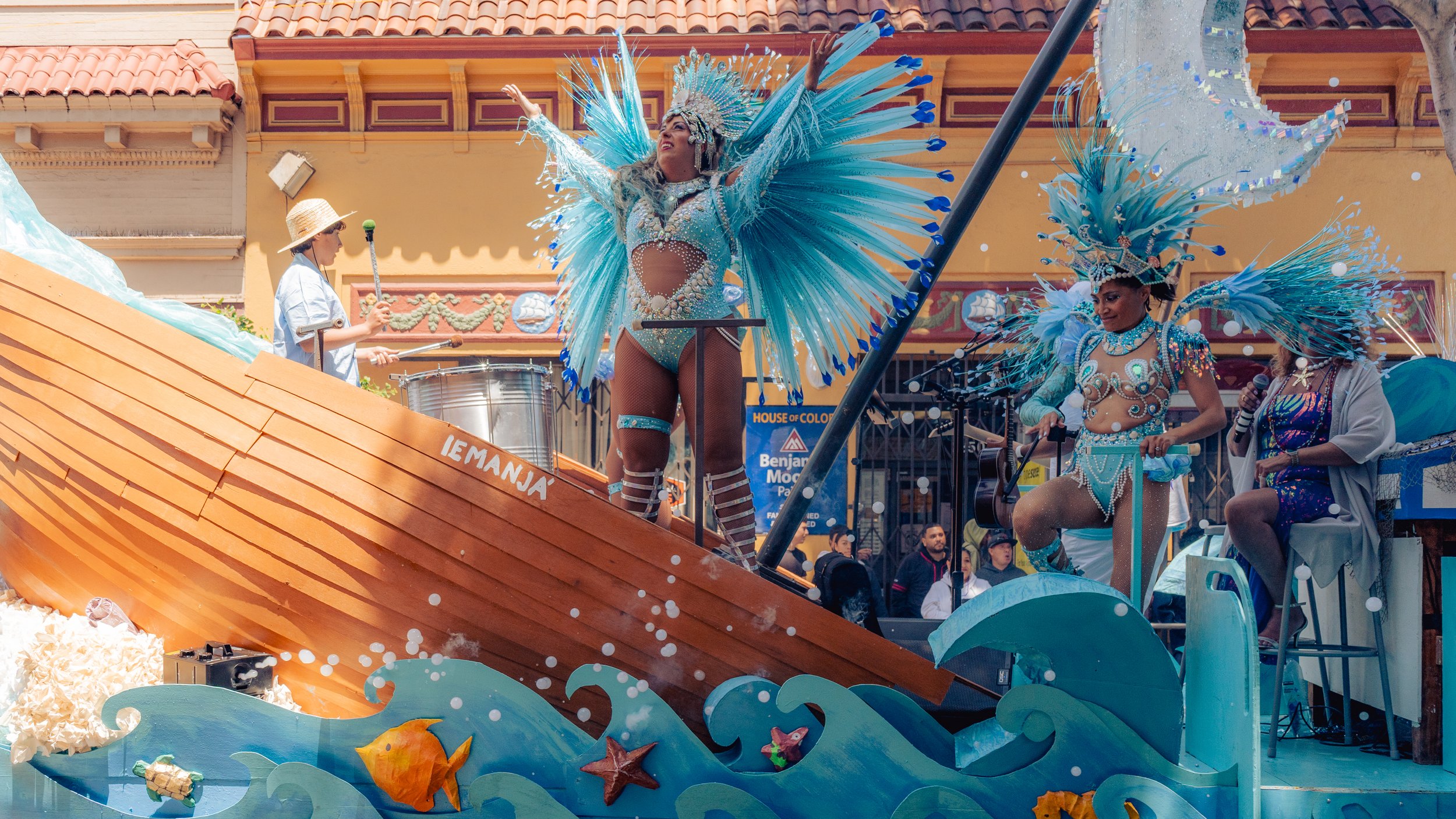 Colorful parade float with women in elaborate, feathered costumes performing, with a musician playing drums nearby, and onlookers in the background.
