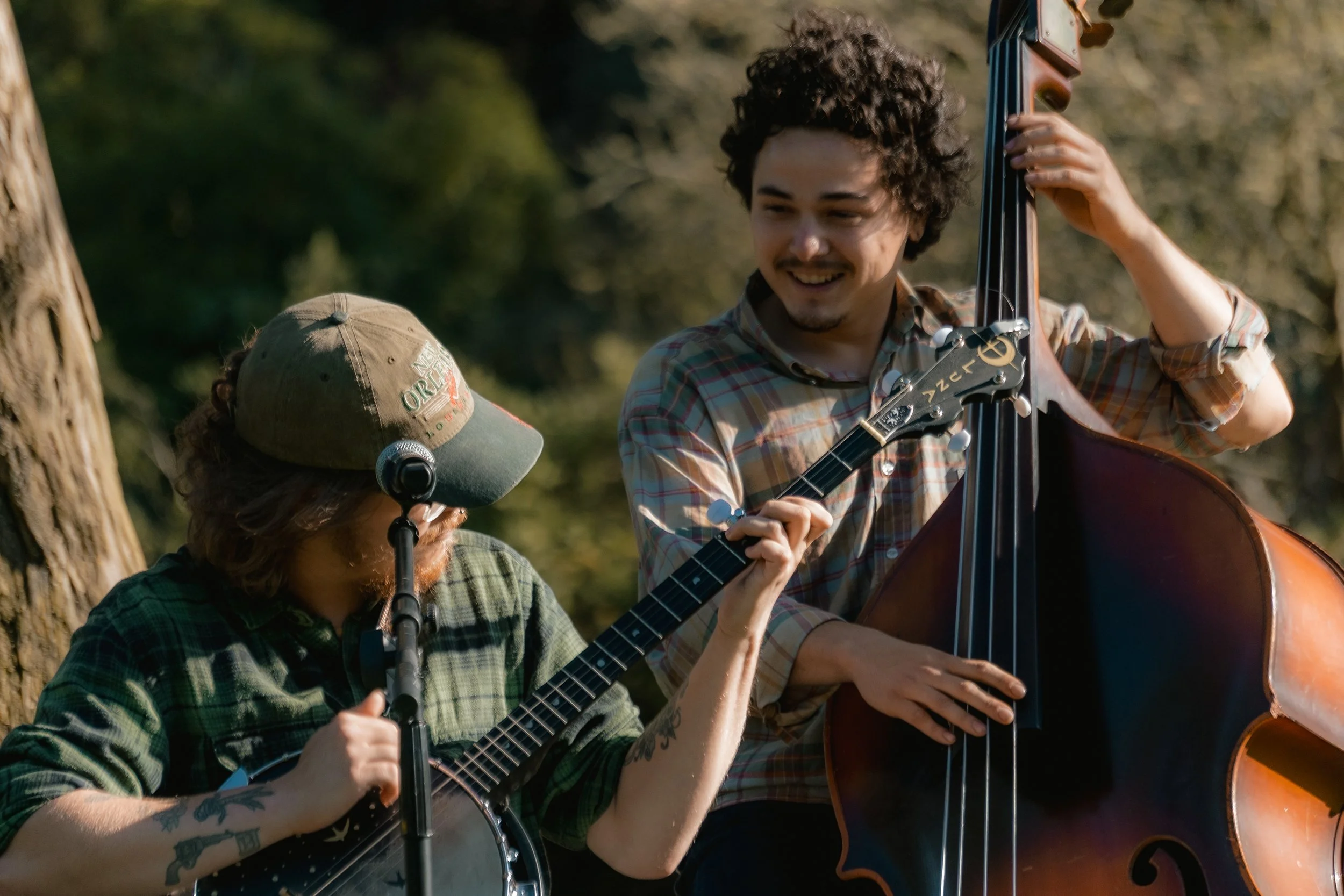 Two men playing string instruments outdoors, one with a banjo and the other with a double bass, smiling and enjoying music together in a natural setting.
