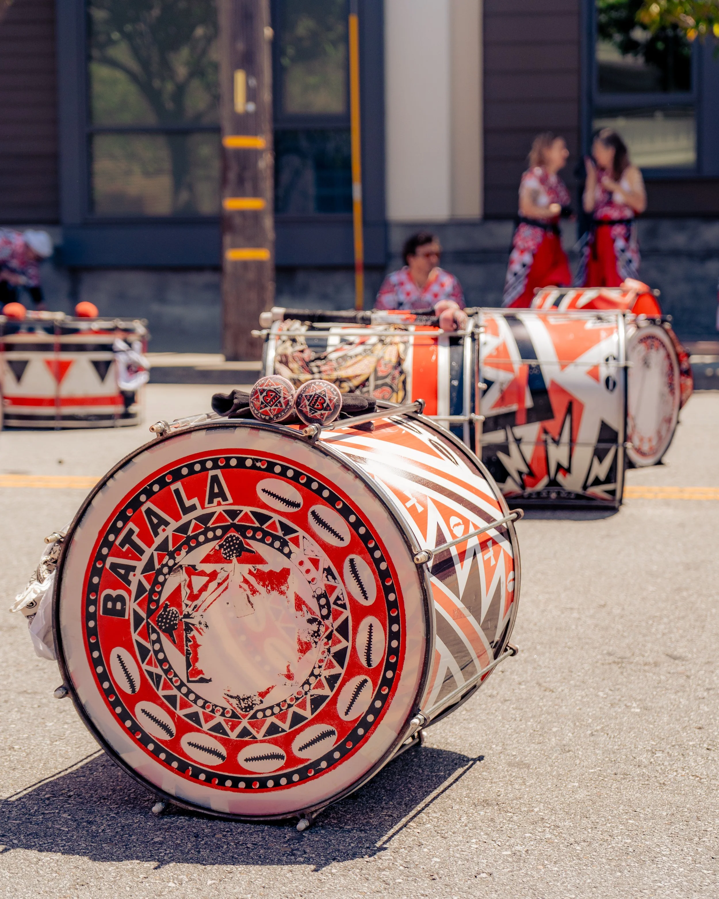 Colorful drums with geometric patterns and the word 'BATALA' on the front, placed on a street during a parade or festival, with people in traditional attire in the background.