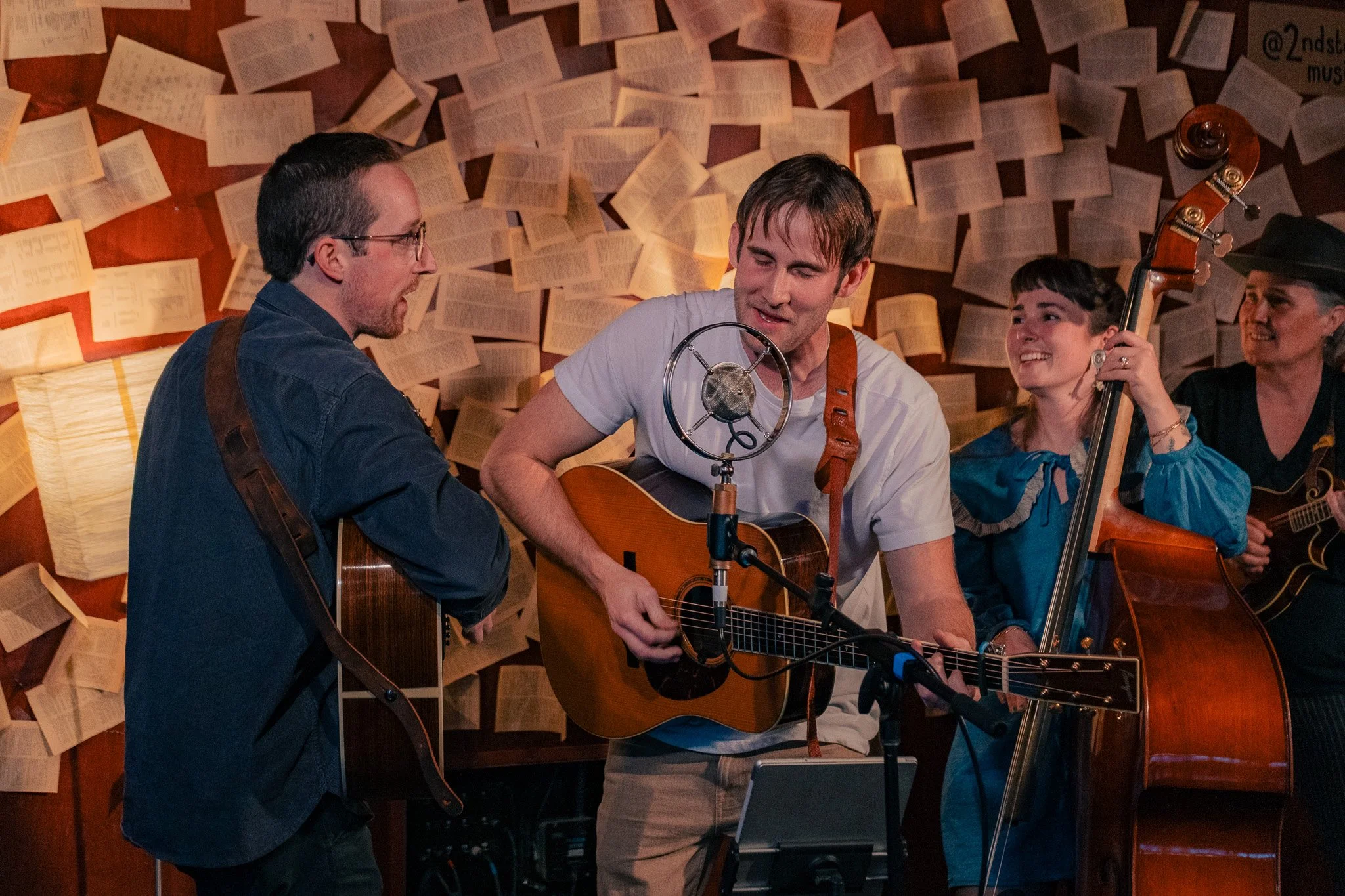 Four musicians performing, two playing guitars and one playing a double bass, with a backdrop of floating pages or sheets of paper.