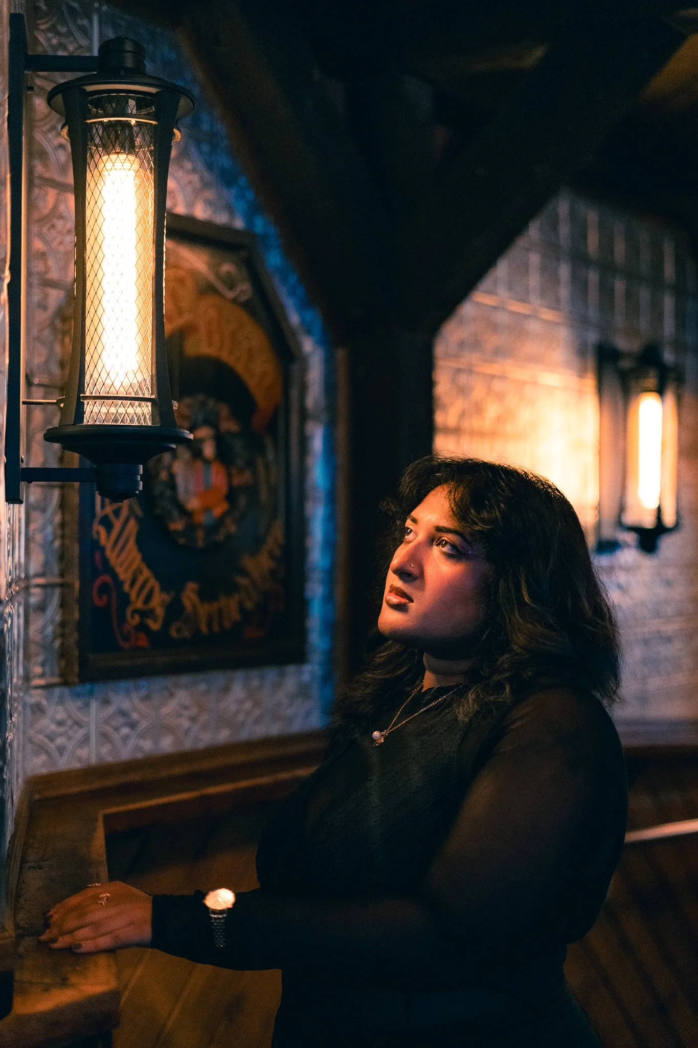 A woman with dark wavy hair and a nose piercing looks thoughtfully at a warm light fixture on a brick wall in a dimly lit, cozy room.