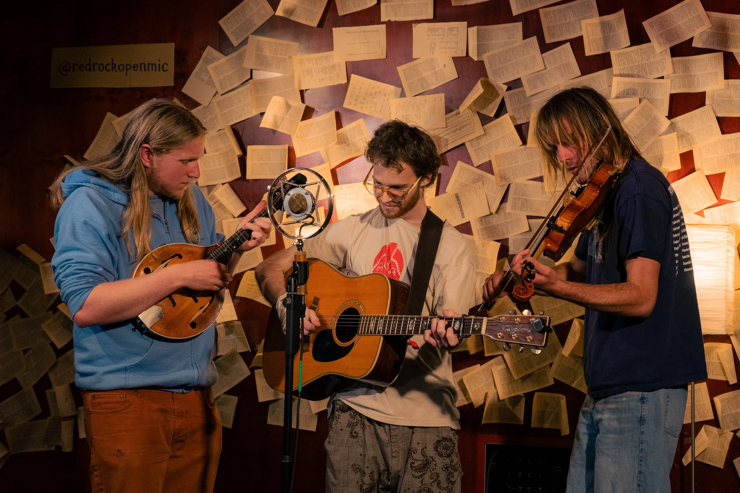 Three musicians playing instruments in front of a wall covered with open books. The musician on the left is playing a mandolin, the middle one is playing an acoustic guitar, and the right one is playing a violin. A microphone is positioned in front o