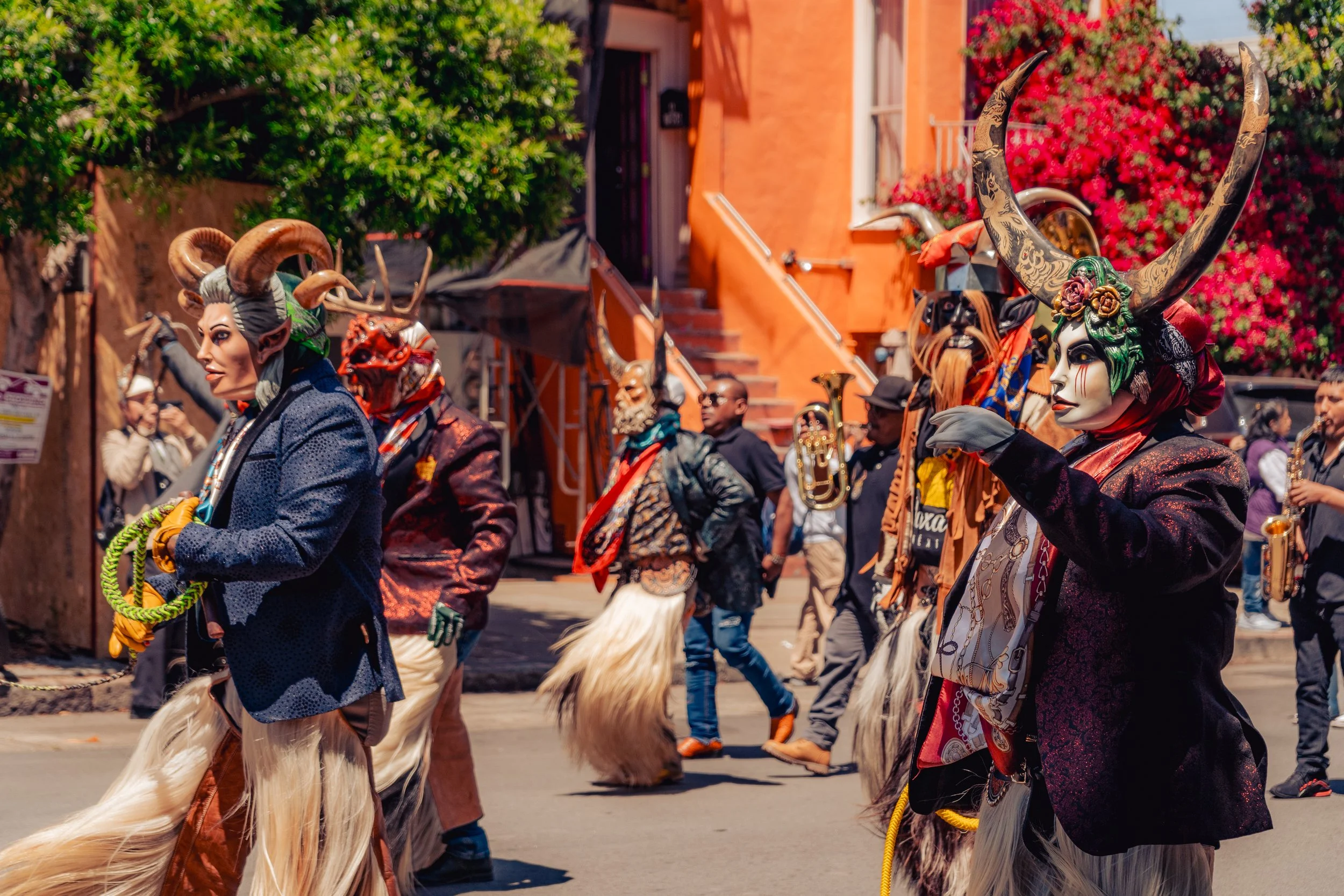 People in colorful costumes and masks with horns, furry legs, and musical instruments participate in a street parade with vibrant buildings and blooming flowers in the background.