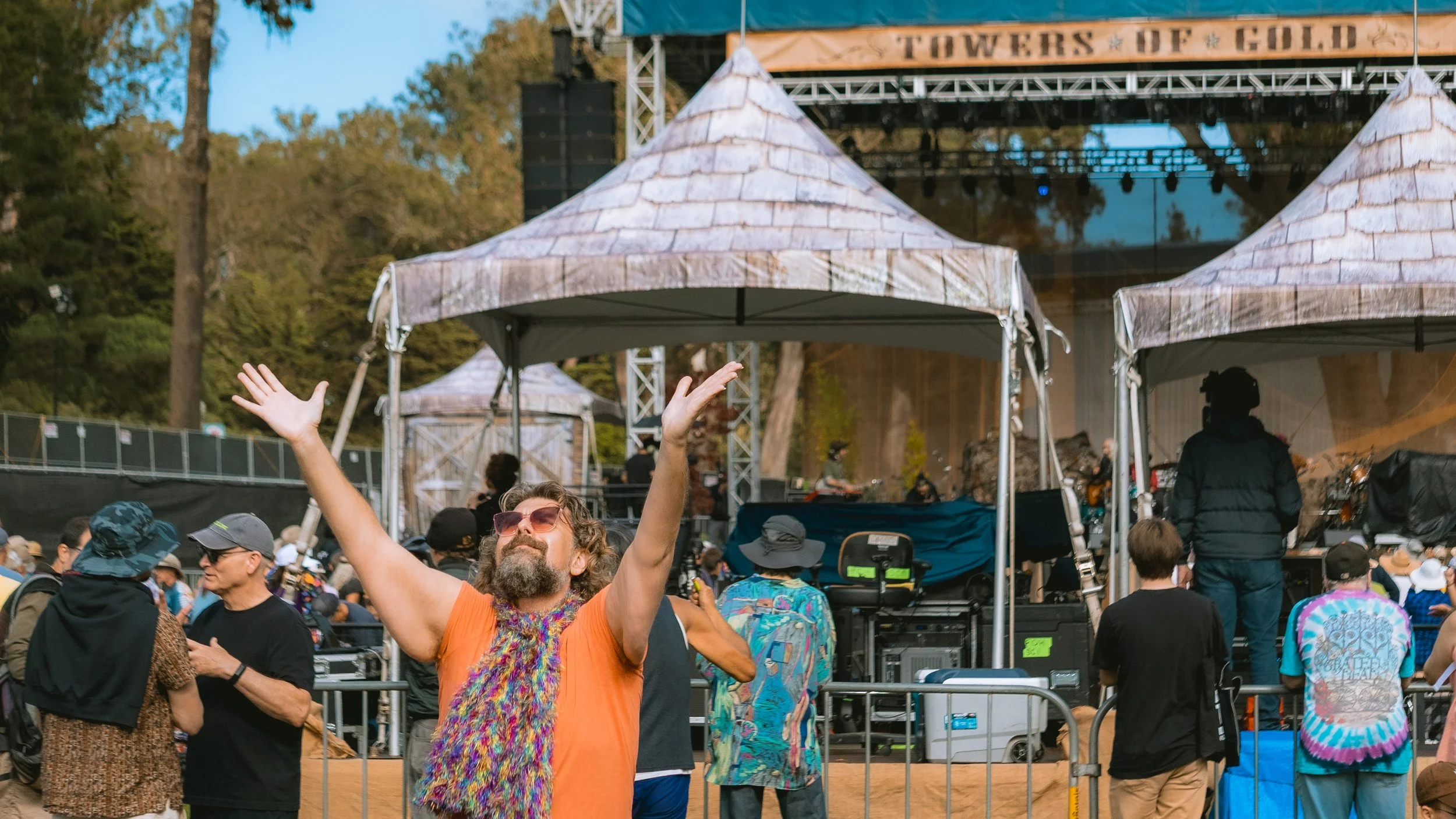 A man with sunglasses, gray beard, and colorful scarf singing or enjoying a festival outdoors, with a stage and crowd in the background.