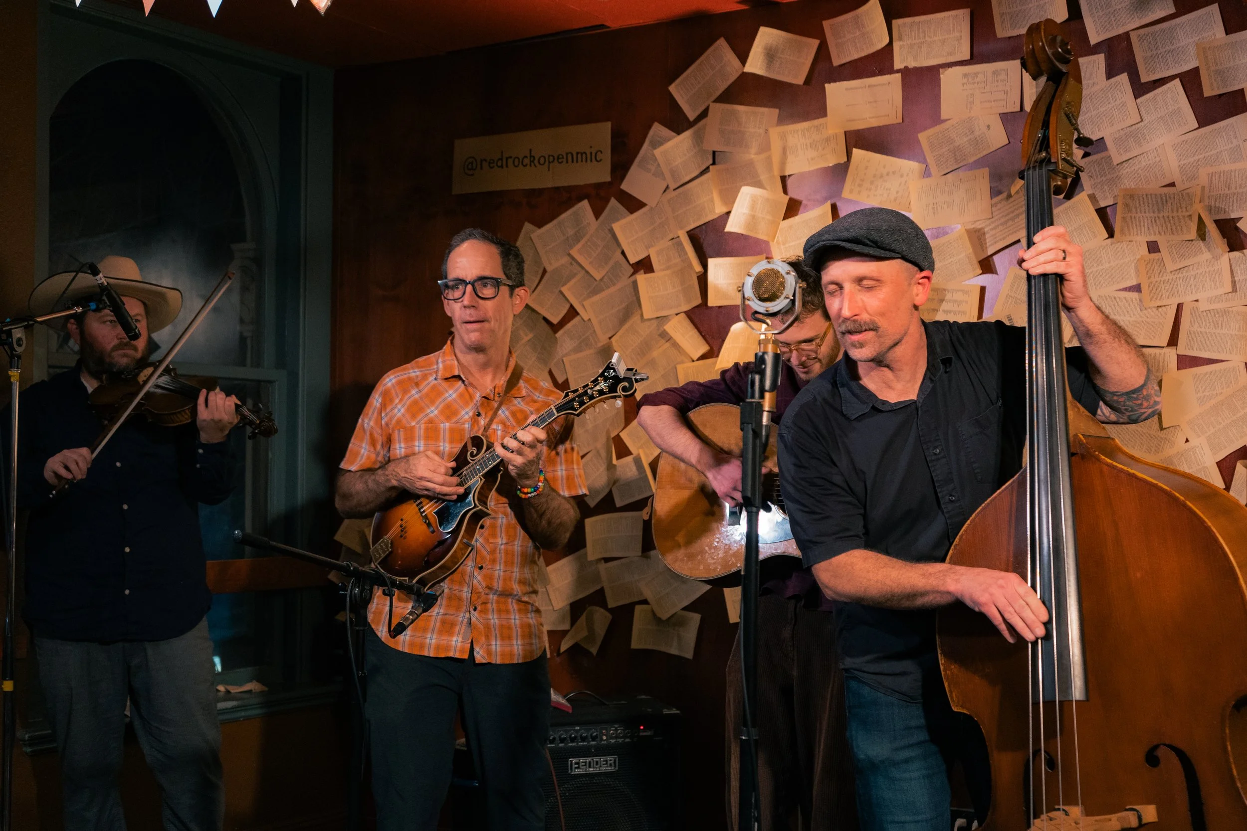 Four musicians performing with string instruments in a cozy, indoor setting with a wall covered in open books and a sign that says '@redrockopenmic'.