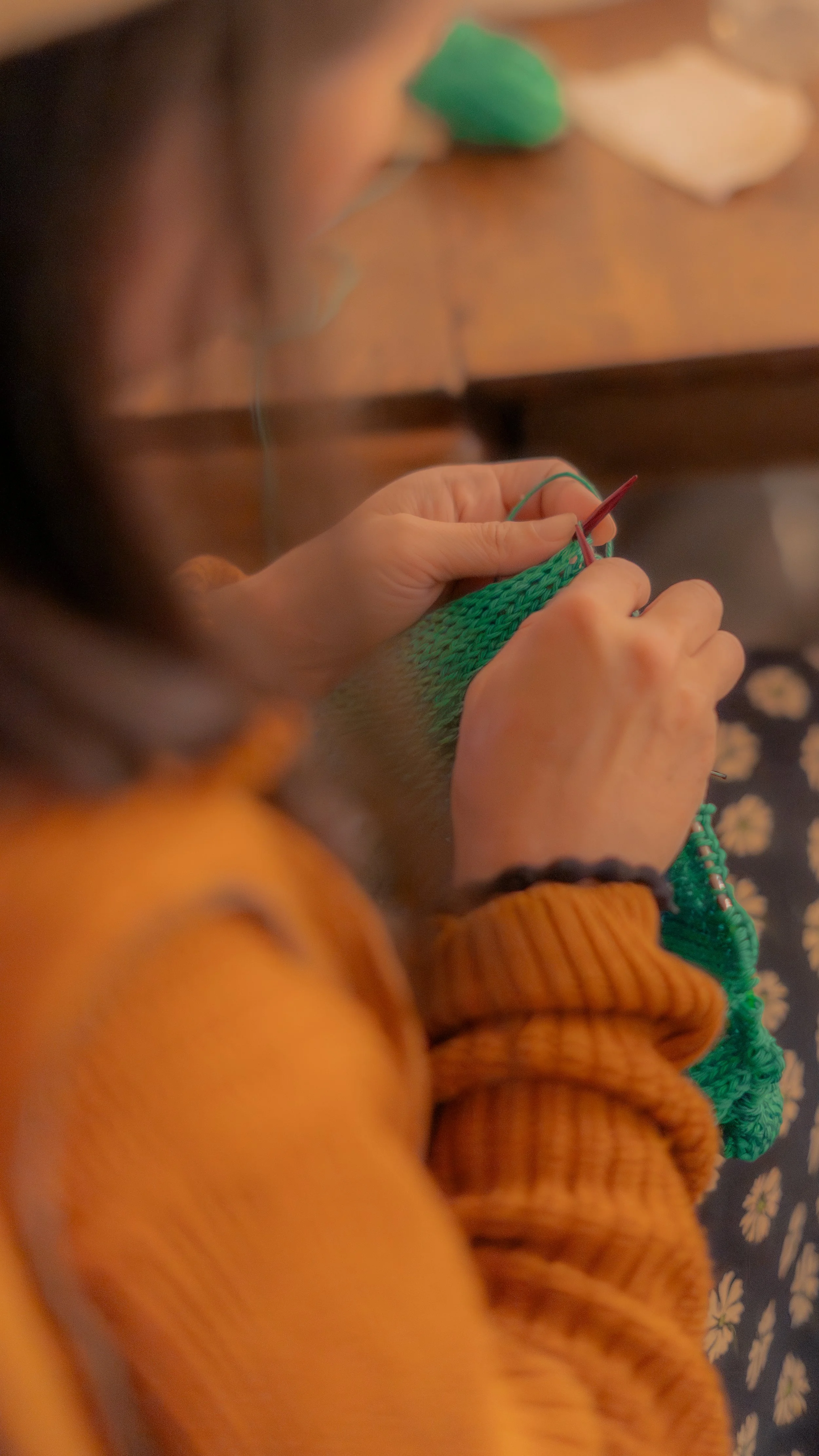 Person knitting with green yarn, sitting at a wooden table with a floral pattern fabric nearby.