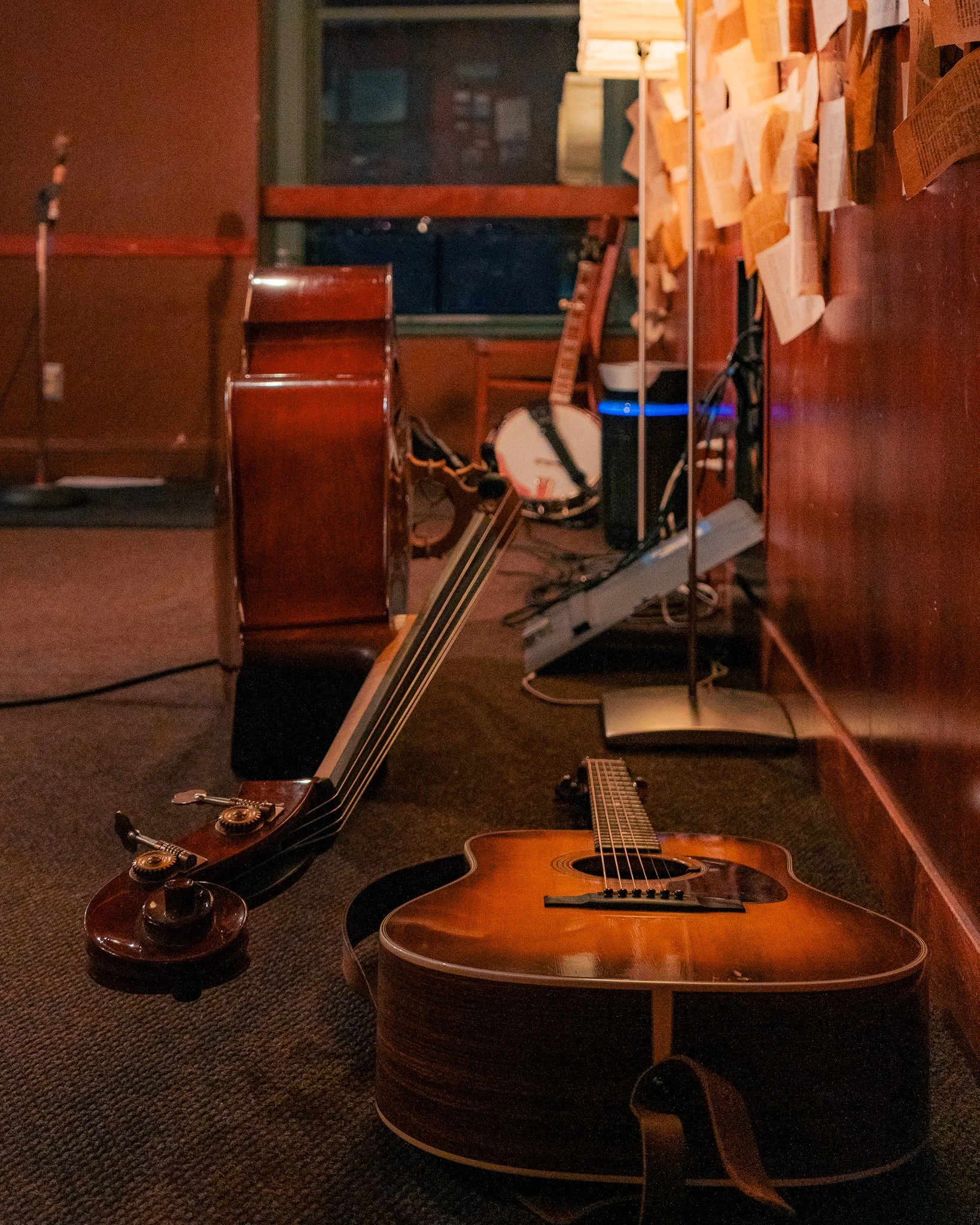 A room with a classical guitar on the floor, an acoustic guitar on the floor, a drum, and various other musical instruments and equipment, with papers stuck to the wall and a window in the background.