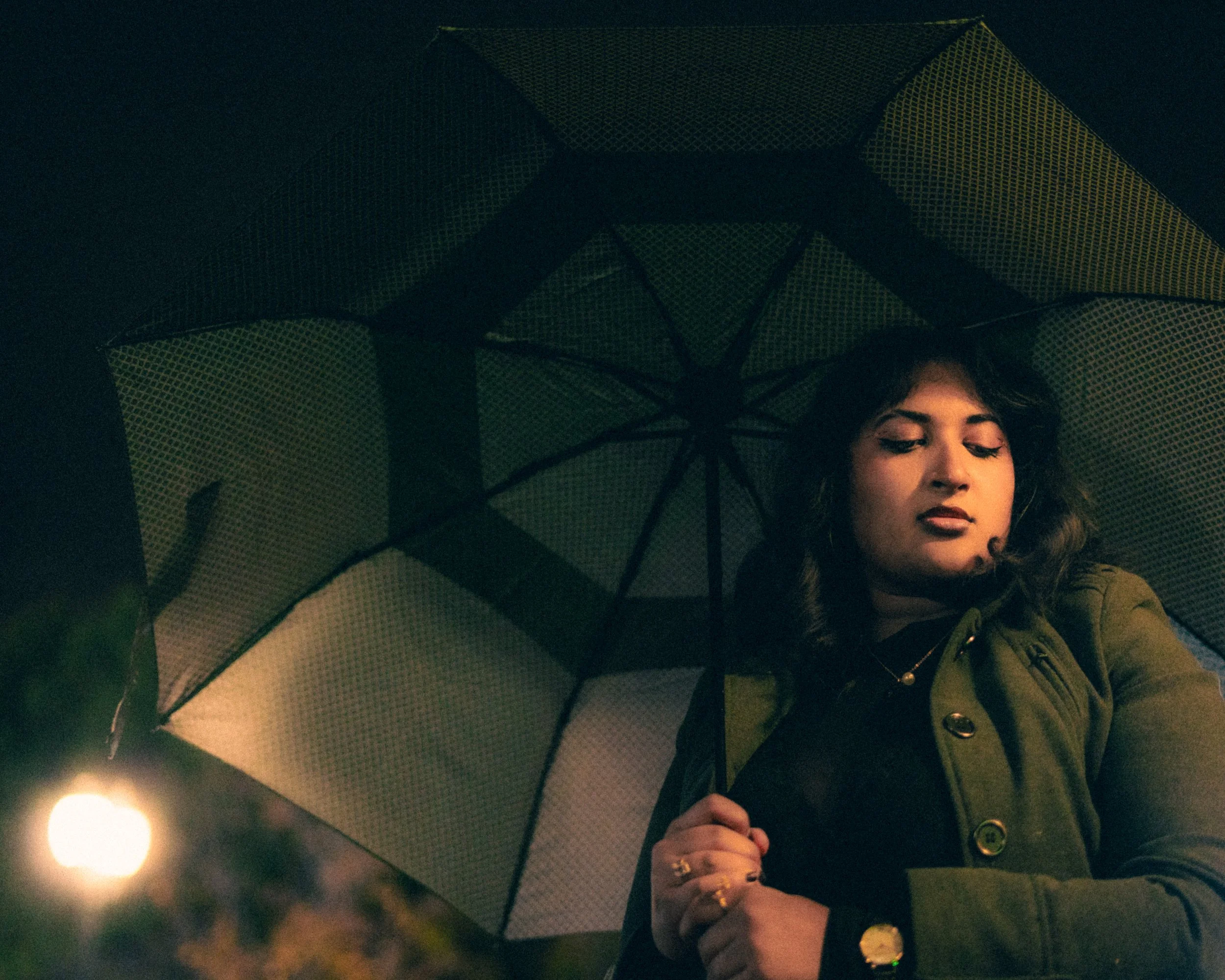 A woman with dark wavy hair holding an umbrella inside a dark room, with a glow of light in the foreground.