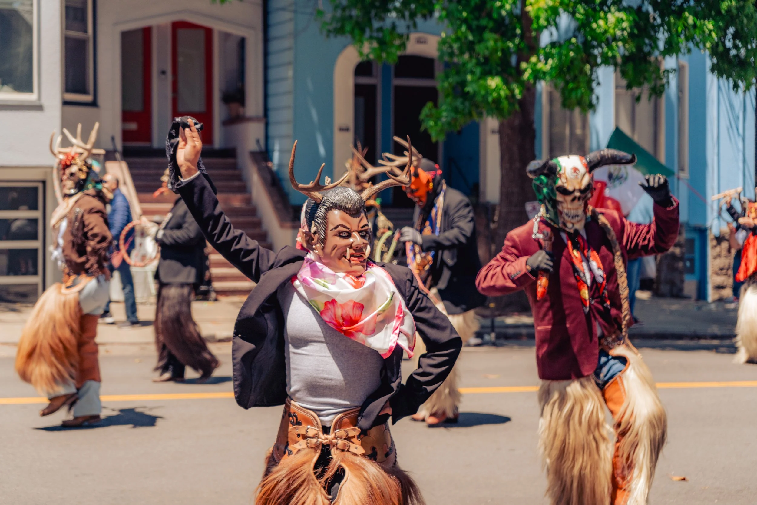 People wearing colorful costumes with masks and horns, participating in a parade or festival on a city street.