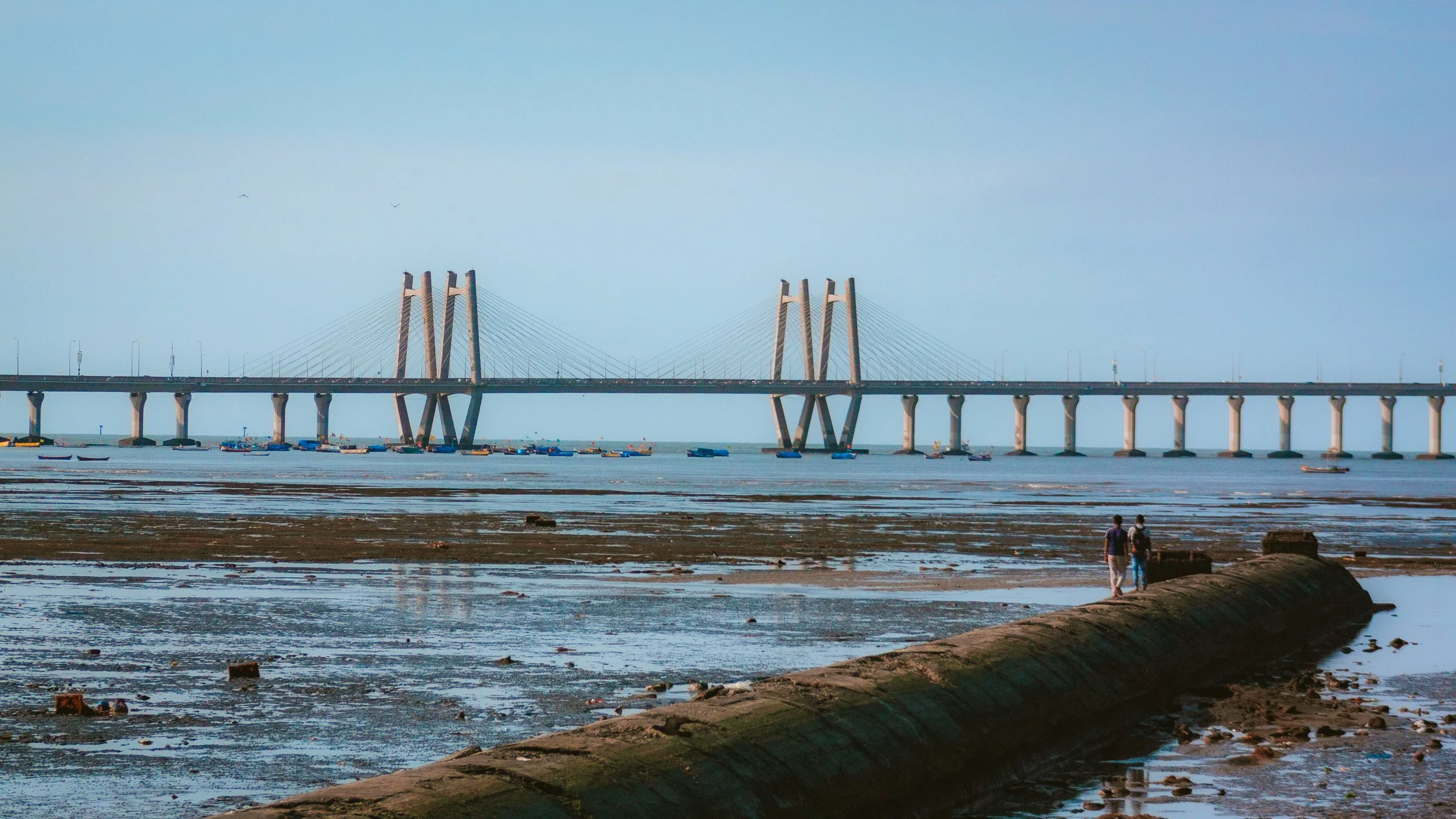 Long bridge with tall support towers and suspension cables spans over a body of water, with boats visible below. Two people walk along a concrete pathway in the foreground on a muddy shoreline during daytime.