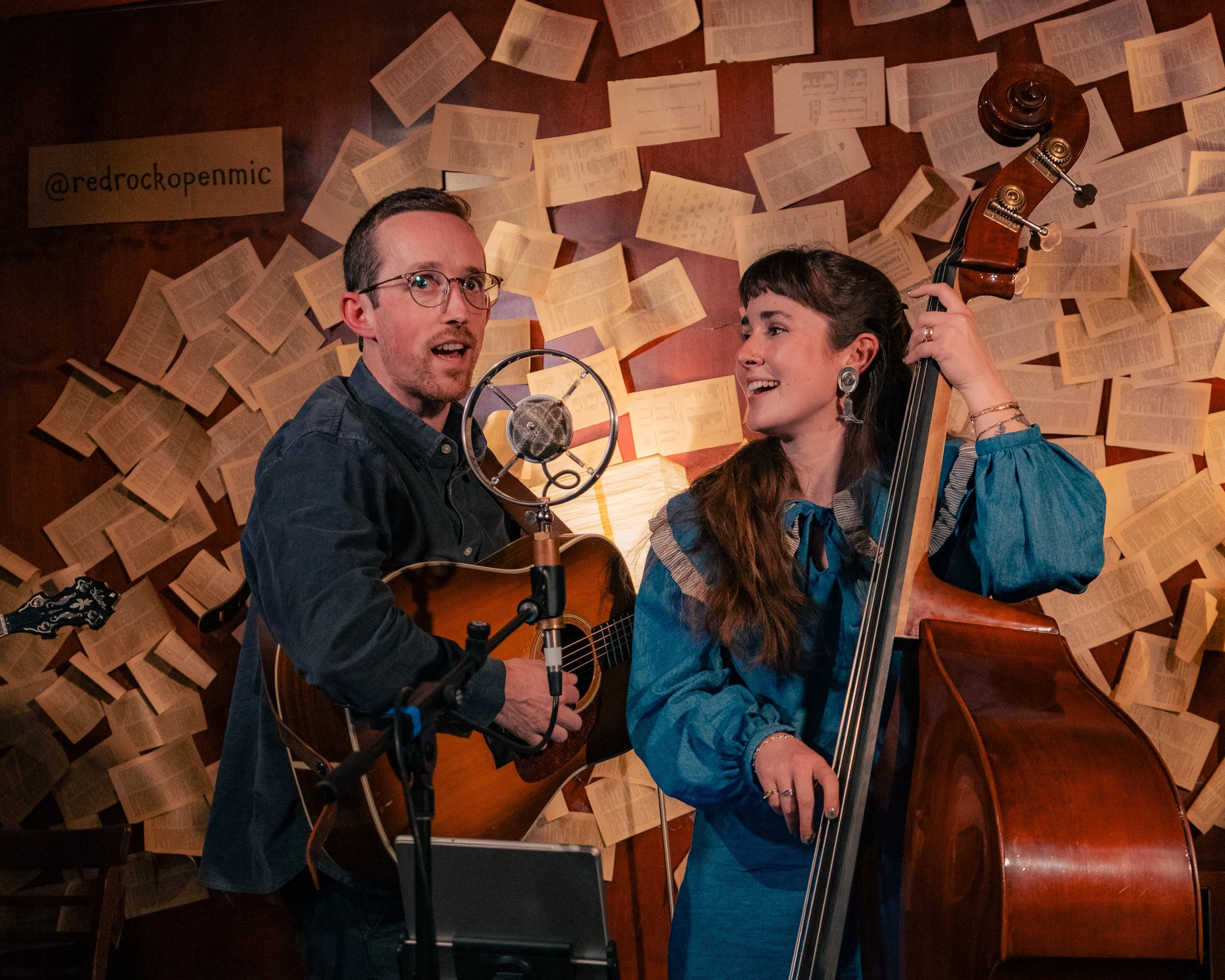 Two musicians, a man playing guitar and a woman playing double bass, perform in front of a wall decorated with pages of open books. A microphone is positioned in front of the man.